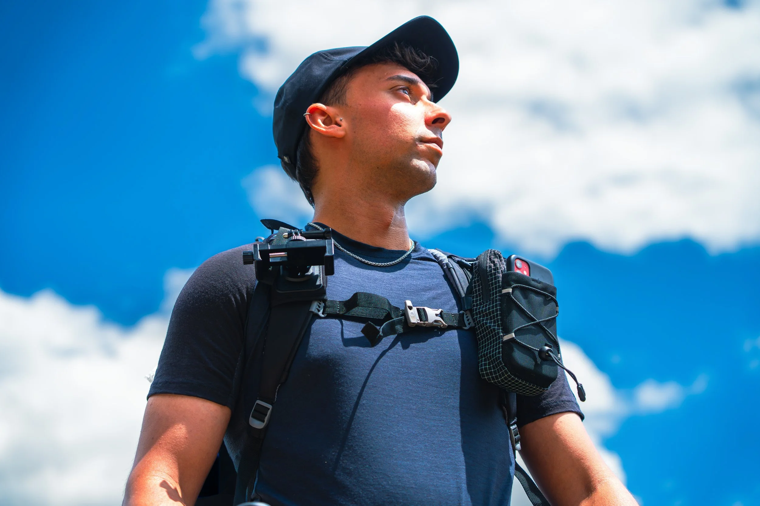 Joven deportista con mochila, gorra y audífonos en un paisaje de montañas y cielo nublado.