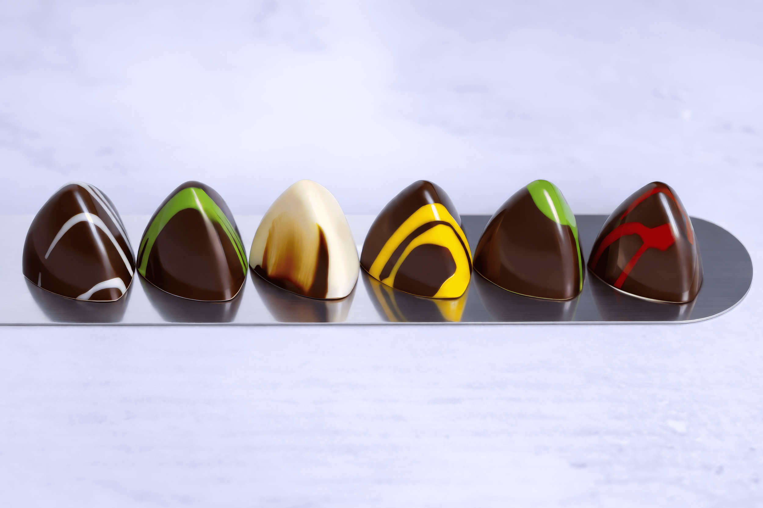 Seis chocolates decorados con glaseado de colores diferentes, colocados en una bandeja metálica en un fondo claro.