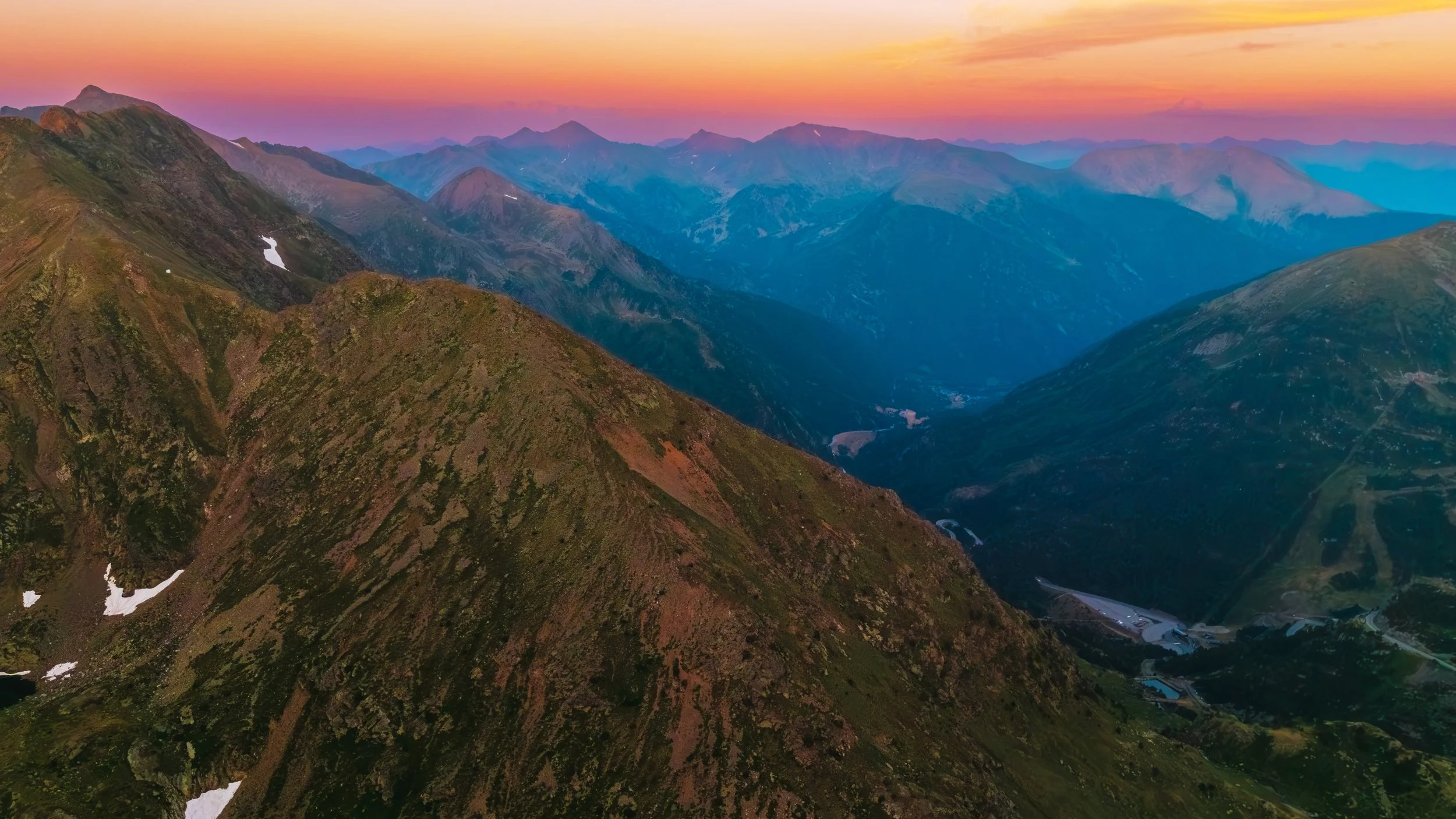 Vista panorámica de montañas durante el atardecer con picos boscosos y algunos restos de nieve en las pendientes.