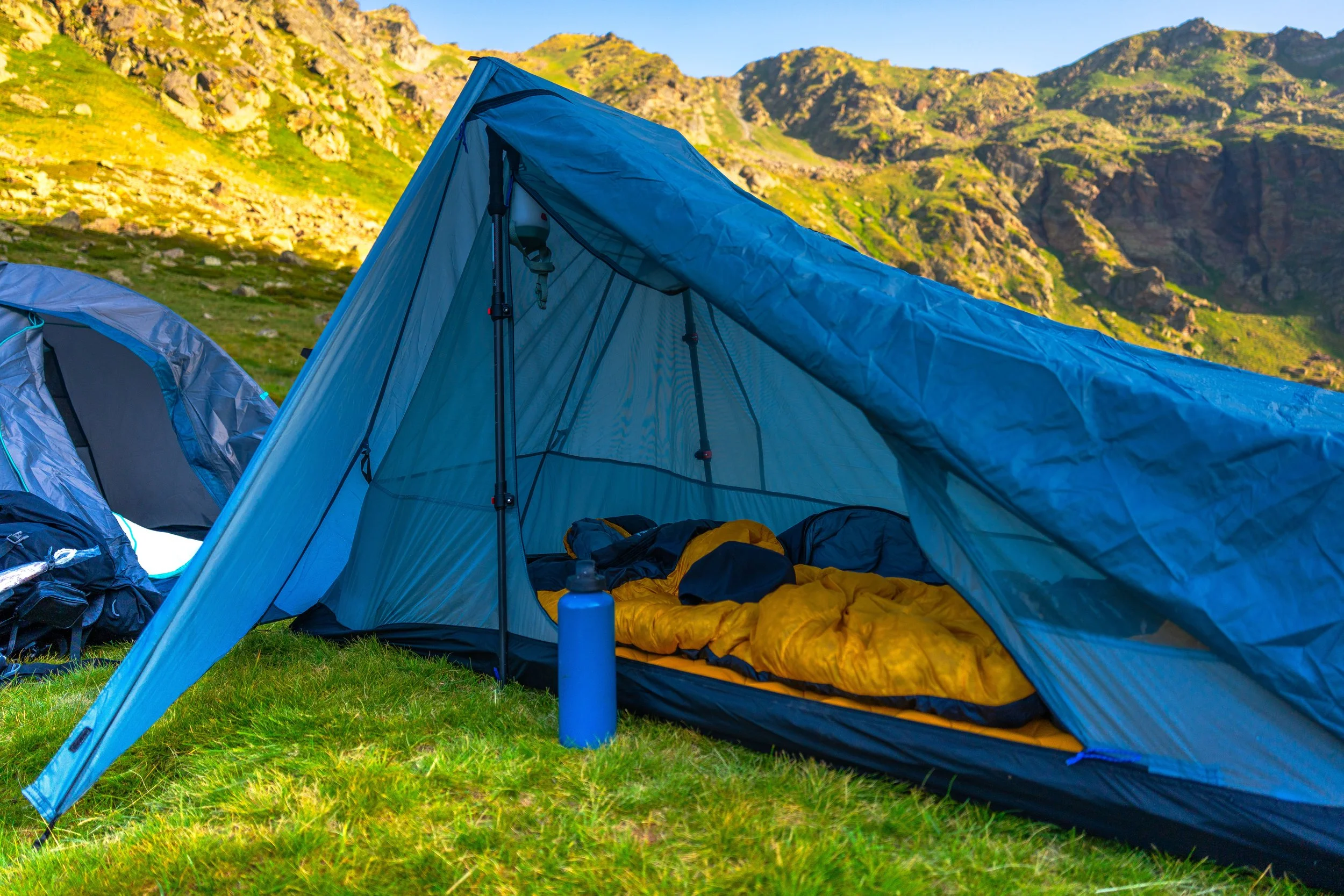 Tienda de campaña azul en un campo con montañas verdes al fondo, con saco de dormir amarillo y otros objetos de camping en el interior.