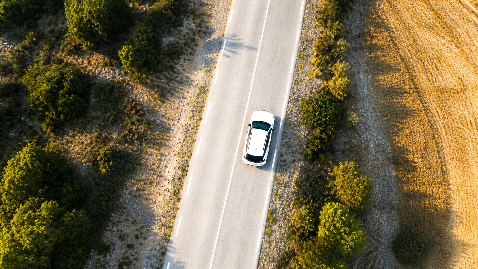 Fotografía aérea de un automóvil blanco conduciendo por una carretera en un área rural rodeada de árboles y campo.