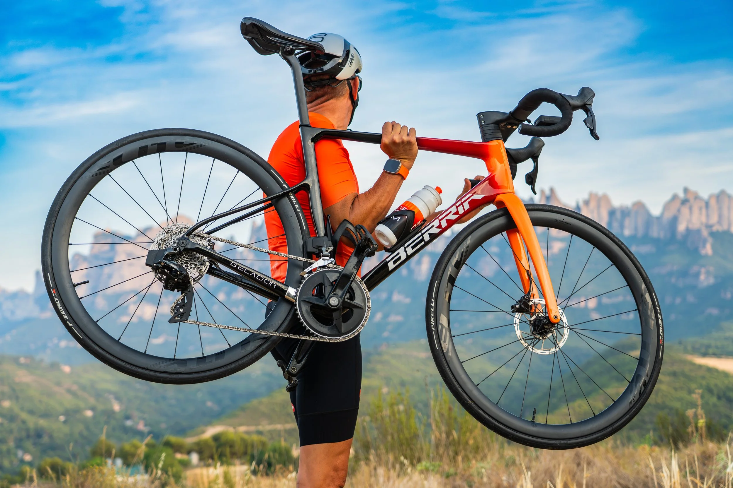 Persona usando un casco y llevando una bicicleta de ruta naranja y negro, en un paisaje con montañas al fondo y cielo despejado.