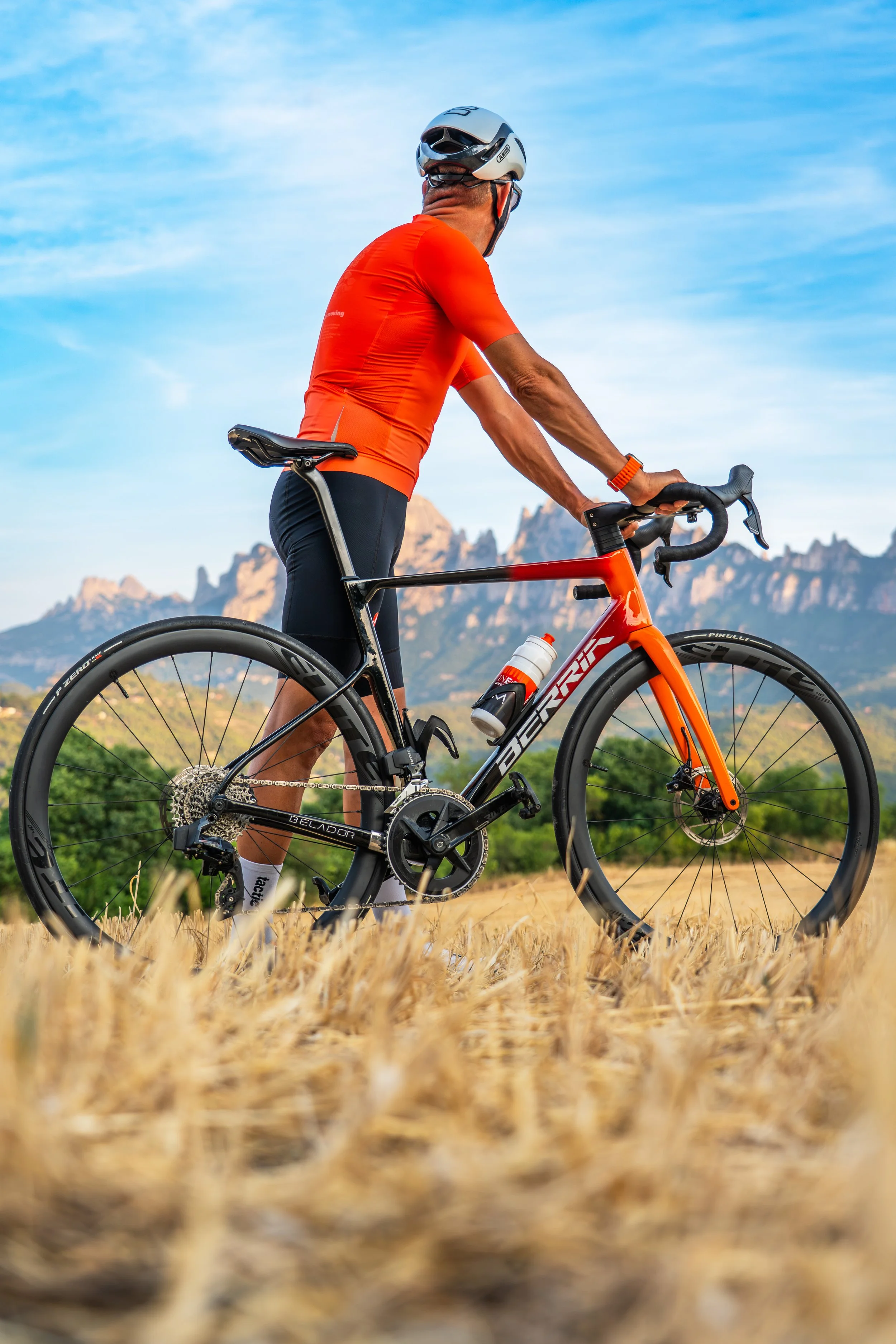 Un ciclista vestido con ropa deportiva naranja, usando casco y gafas, montando una bicicleta de carreras en un campo con césped seco y montañas al fondo bajo un cielo despejado.