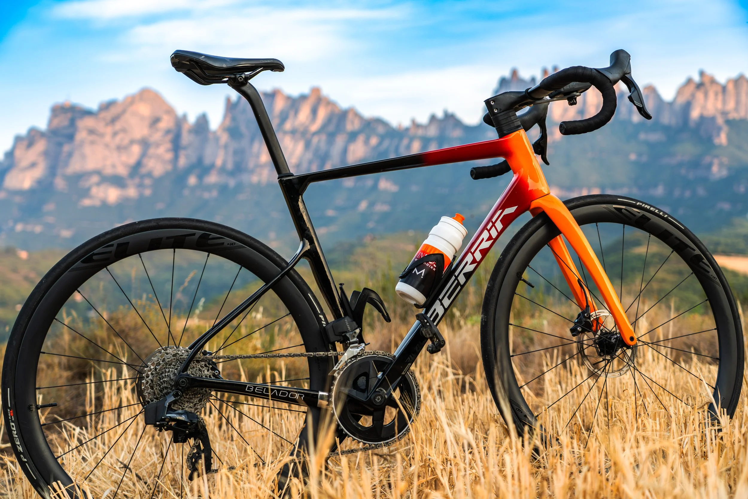 Bicicleta de ruta con cuadro naranja y componentes negros, en un campo de césped seco, con montañas rocosas y cielo azul en el fondo.