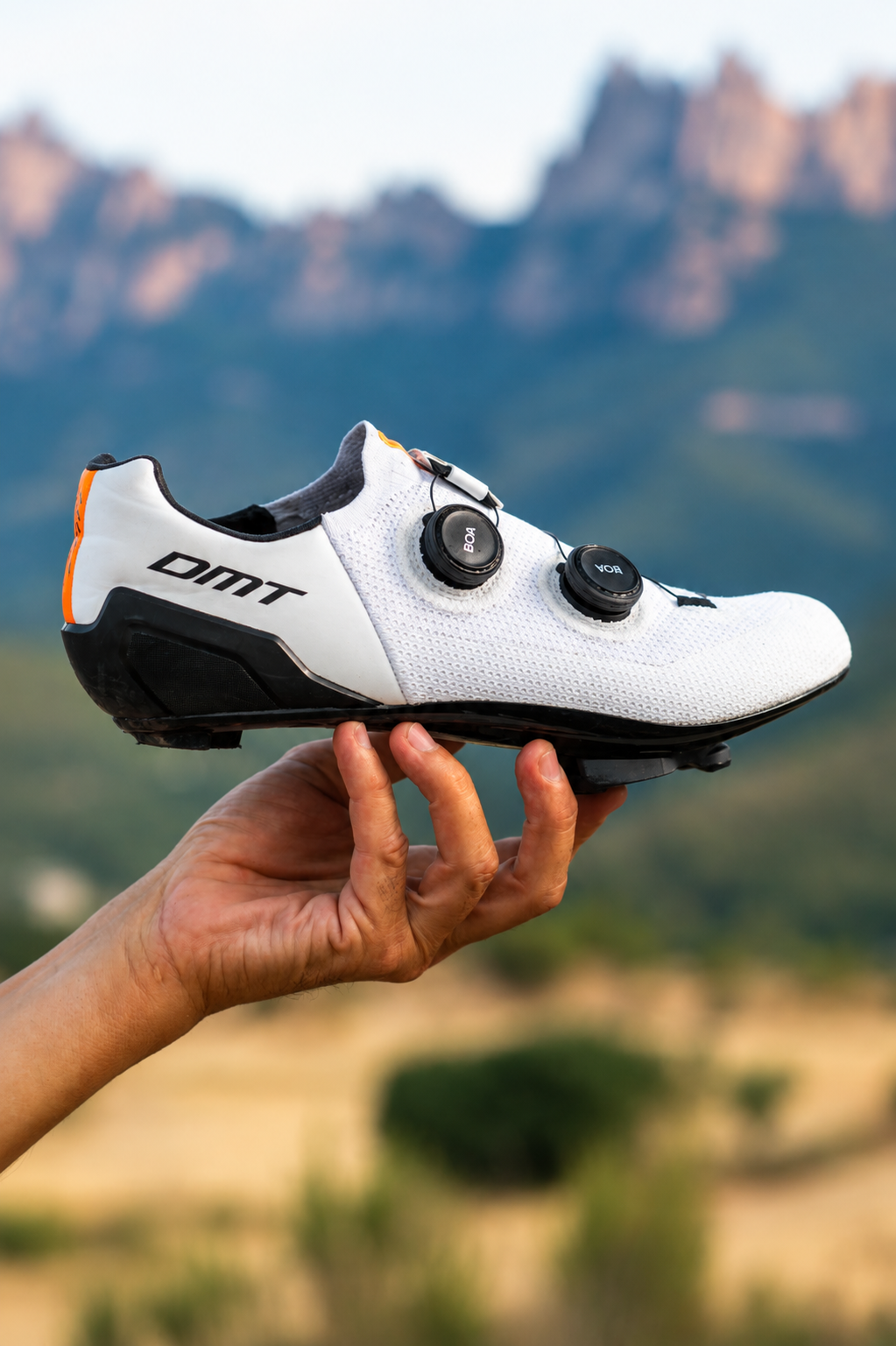 Una mano sostiene una zapatilla de ciclismo con calas, en un fondo de paisaje montañoso borroso.