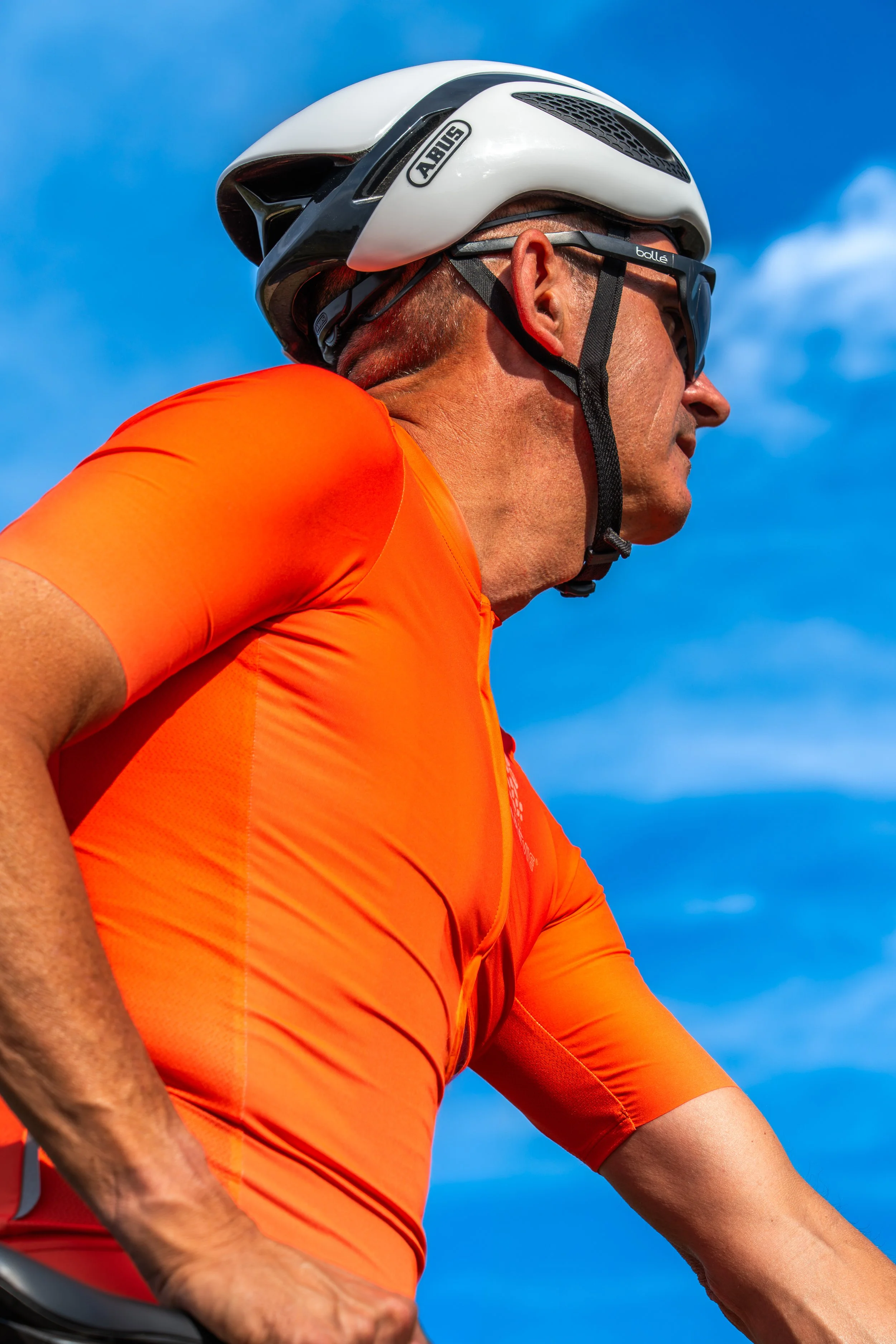 Ciclismo: hombre con casco blanco y gafas de sol, vestido con camiseta naranja, montando bicicleta en cielo despejado.