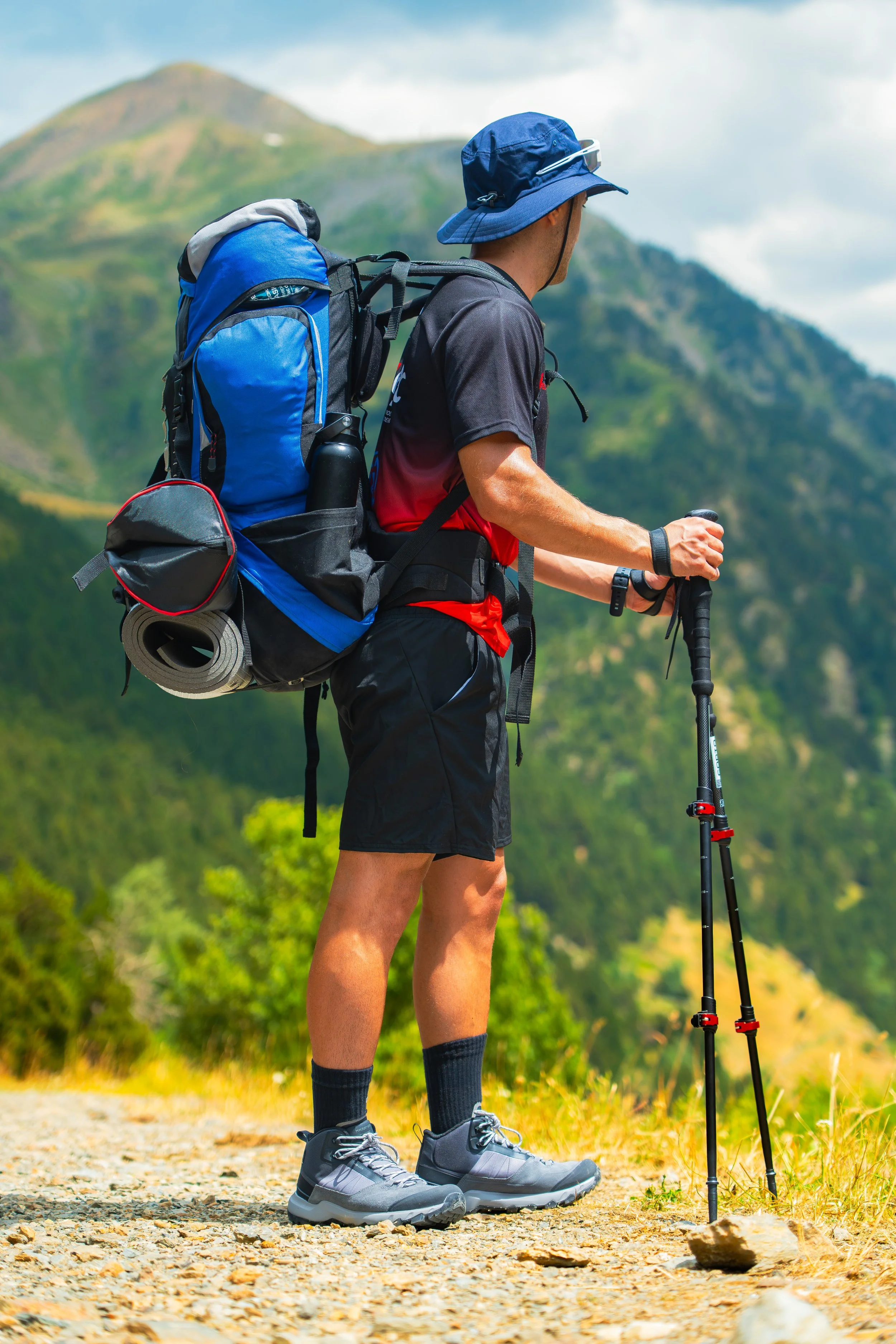 Vendedor usando ropa deportiva, gorra y mochila, caminando con bastón de senderismo en un sendero en las montañas.