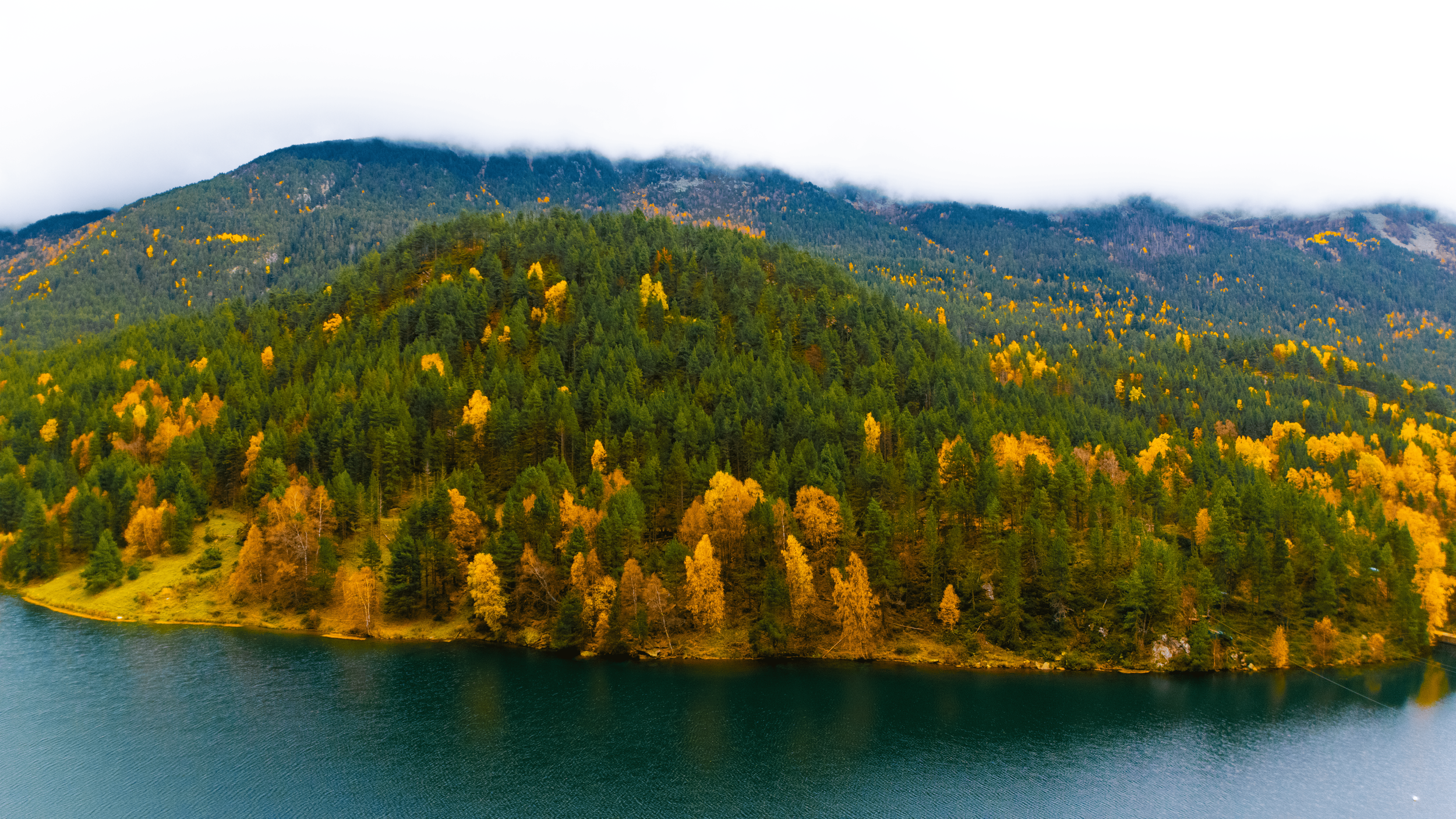 Vista de un lago rodeado de un bosque de árboles verdes y amarillos en otoño, con una montaña en el fondo cubierta parcialmente por nubes.