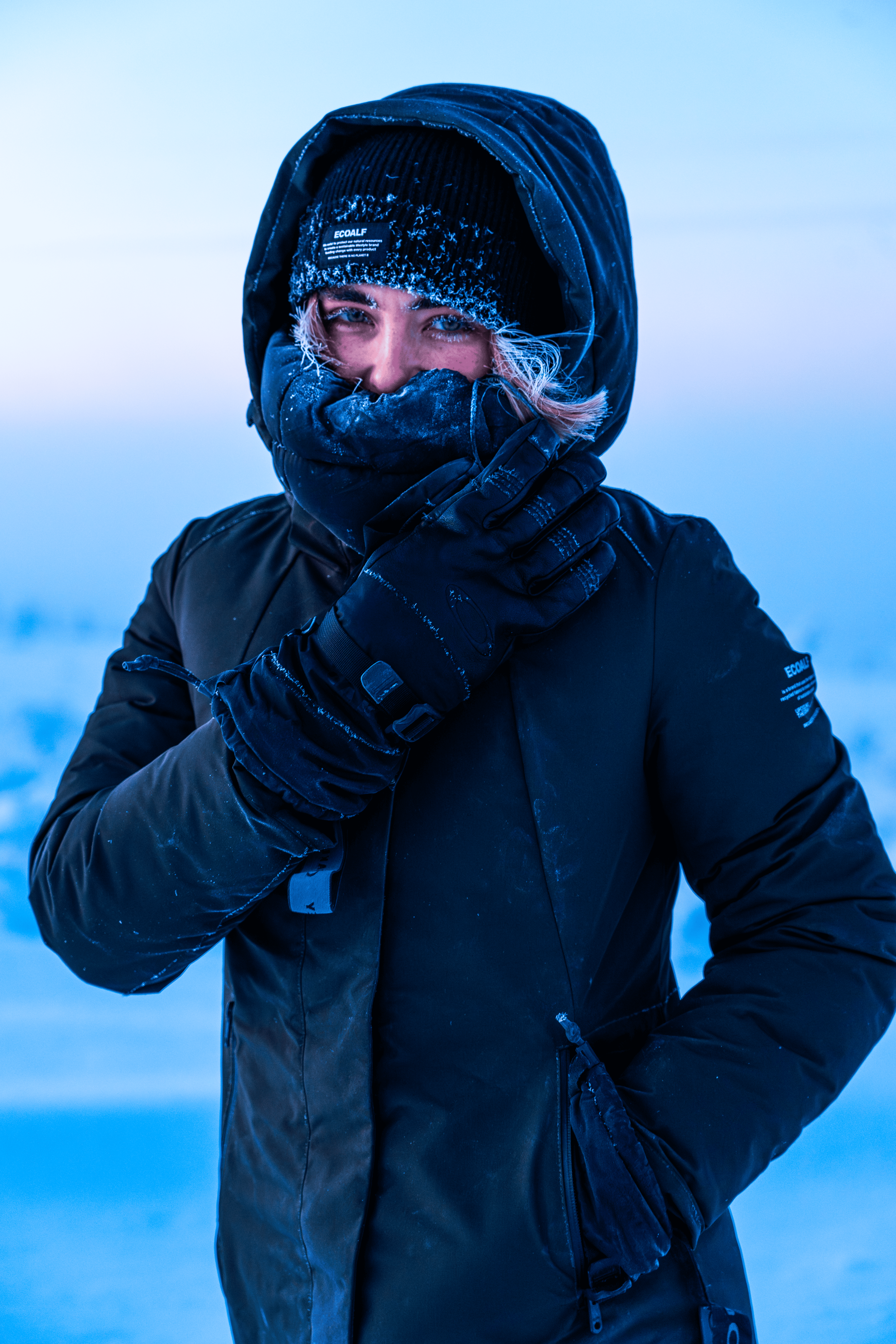 Una persona abrigada con ropa de invierno en un paisaje helado, cubriéndose parcialmente la cara.