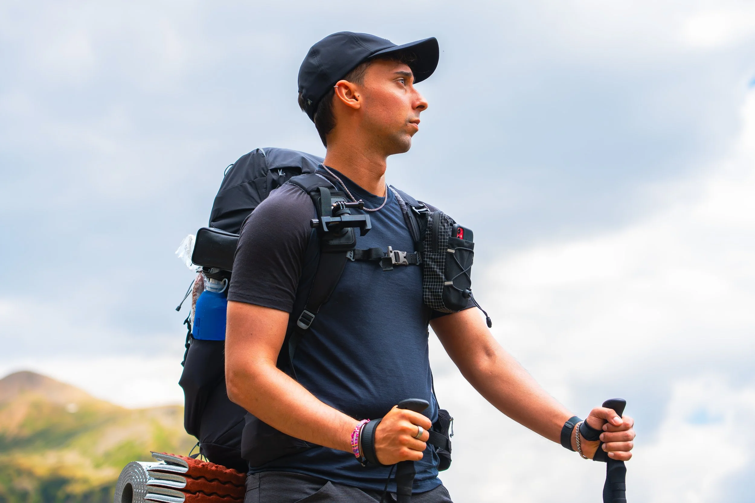 Joven en perfil caminando con mochila en un paisaje natural, usando gorra y ropa cómoda para senderismo.
