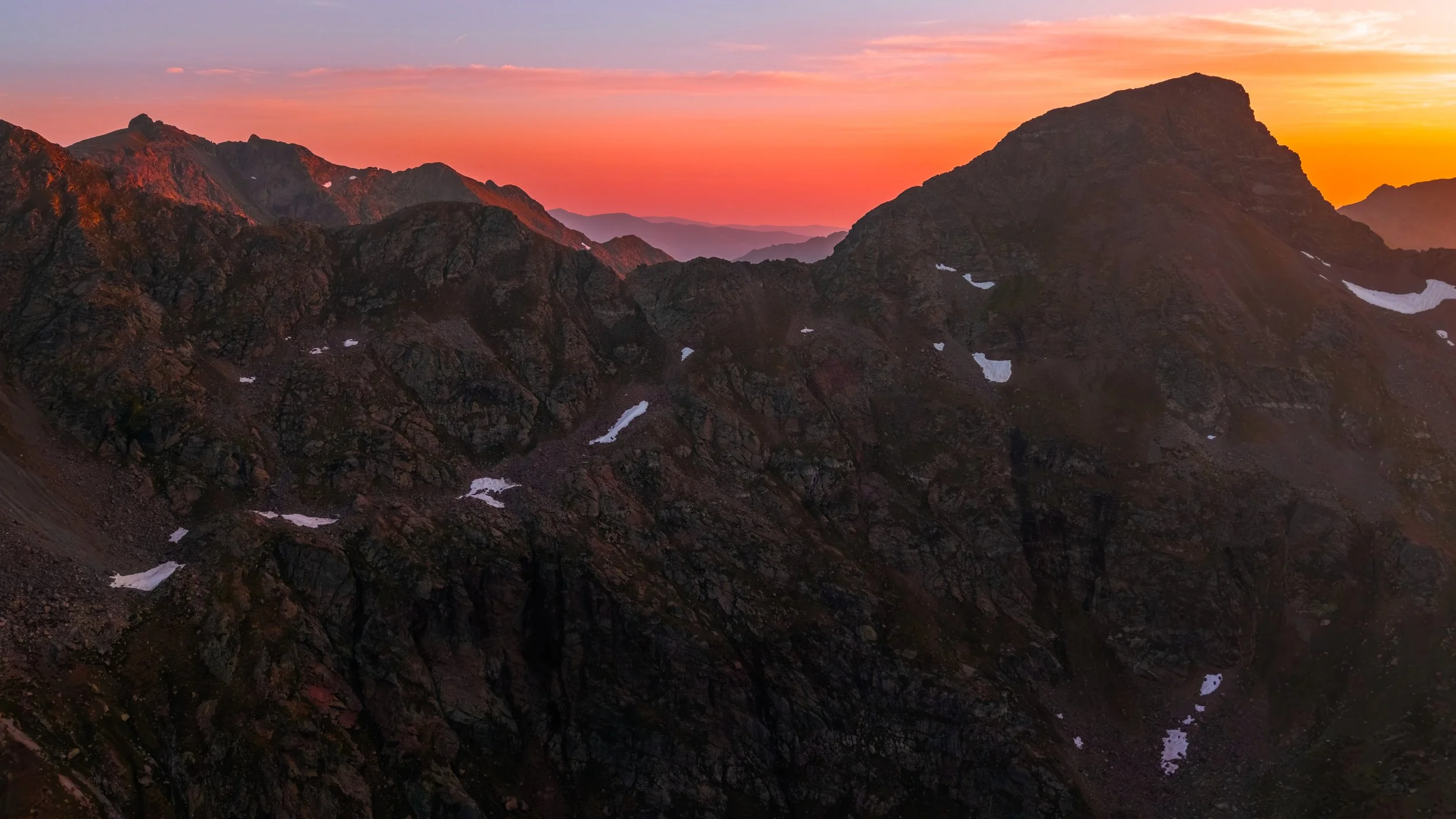 Montañas rocosas durante el atardecer con cielos en tonos rosados y anaranjados, con algunas manchas de nieve en las pendientes.