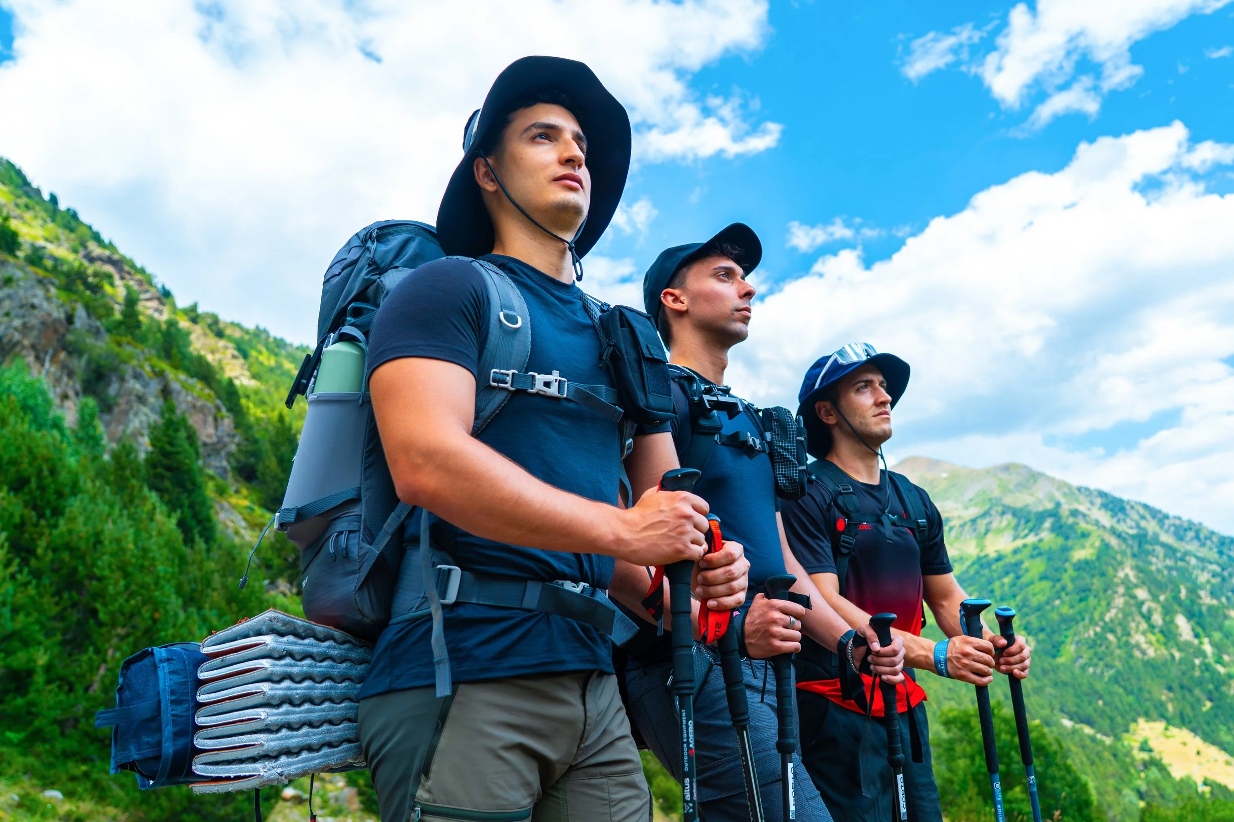 Tres hombres con mochilas y bastones de senderismo observando un paisaje de montañas verdes bajo un cielo con nubes