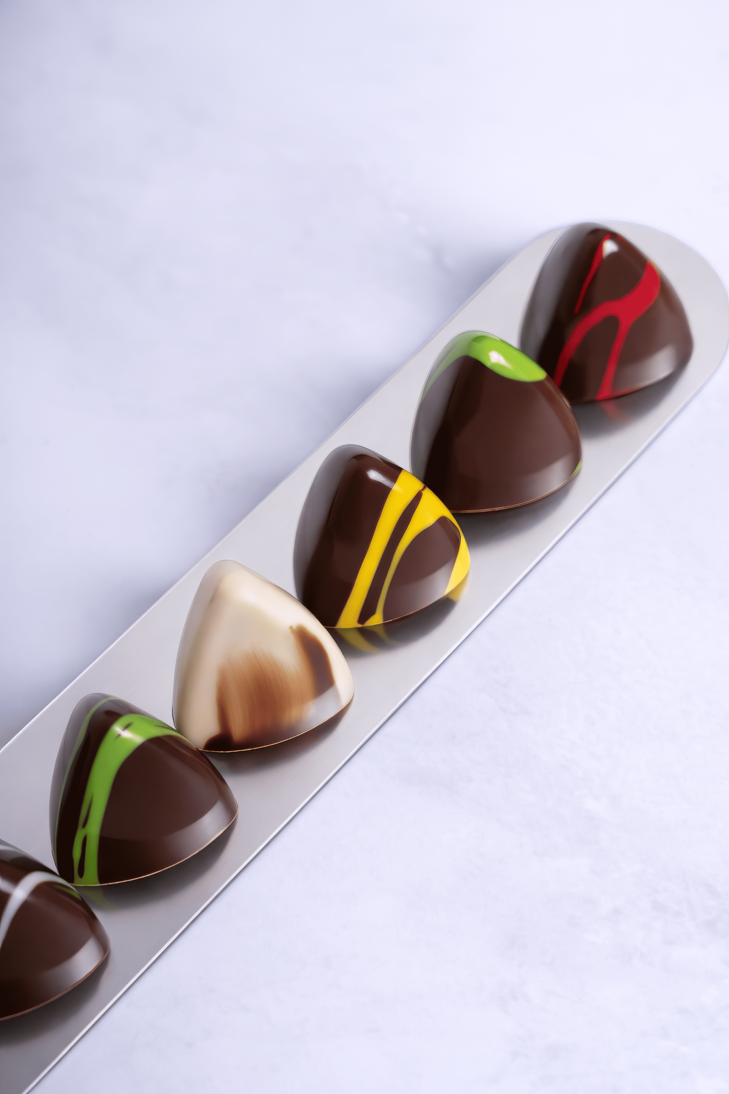 Selección de chocolates de forma ovalada con decoraciones de colores en una bandeja blanca sobre fondo blanco.