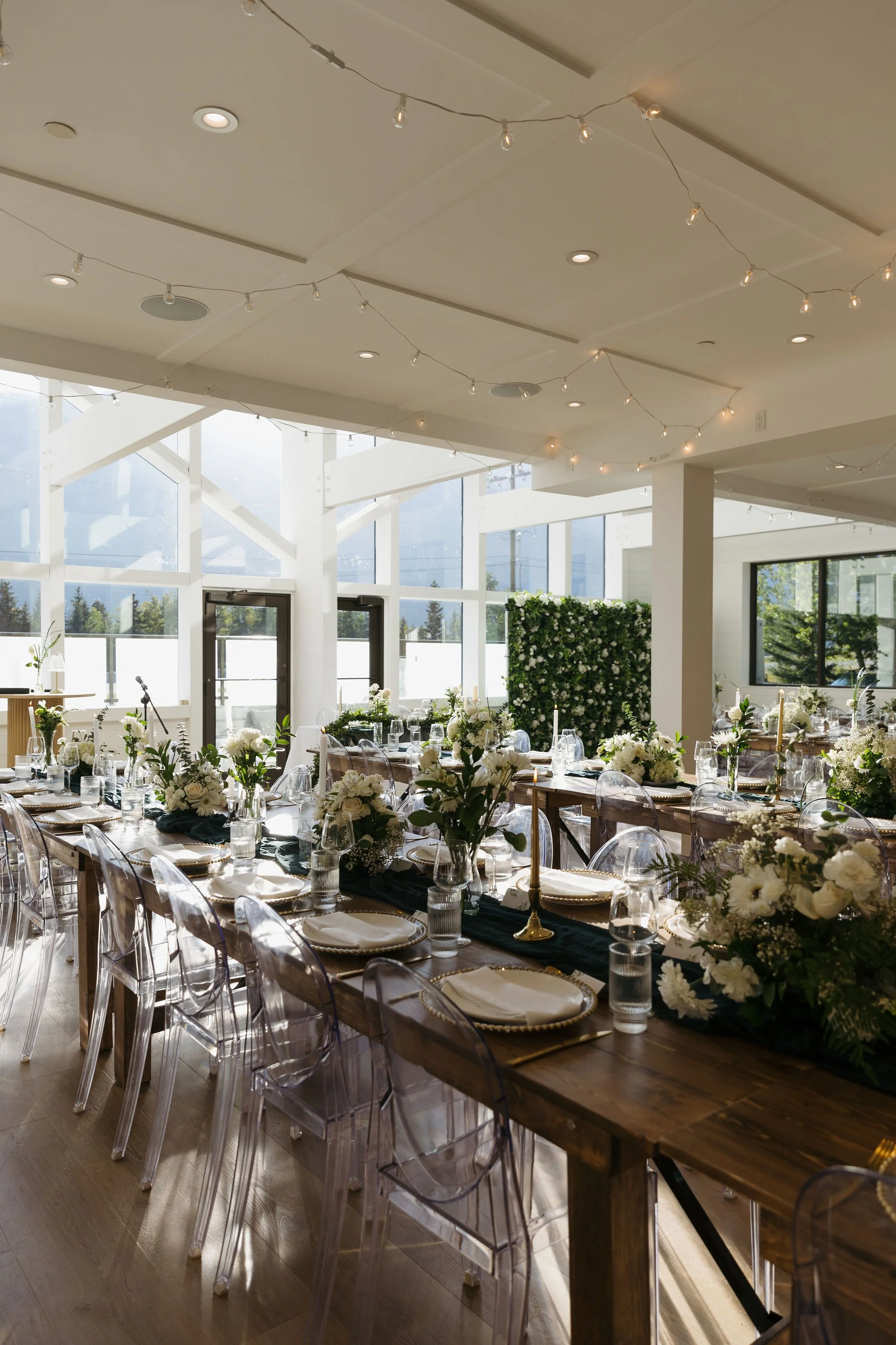 Romantic wedding venue in the Canadian Rockies with bistro string lights, high white ceilings, and panoramic windows. Sophisticated table arrangements with greenery and floral centerpieces complete the look.