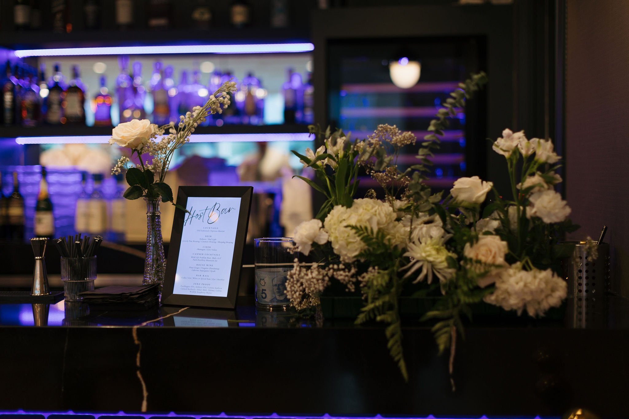 Modern wedding bar setup with custom drink menu, white floral arrangements, and glowing purple backlighting. The perfect stylish cocktail hour space at this contemporary Rocky Mountain venue.