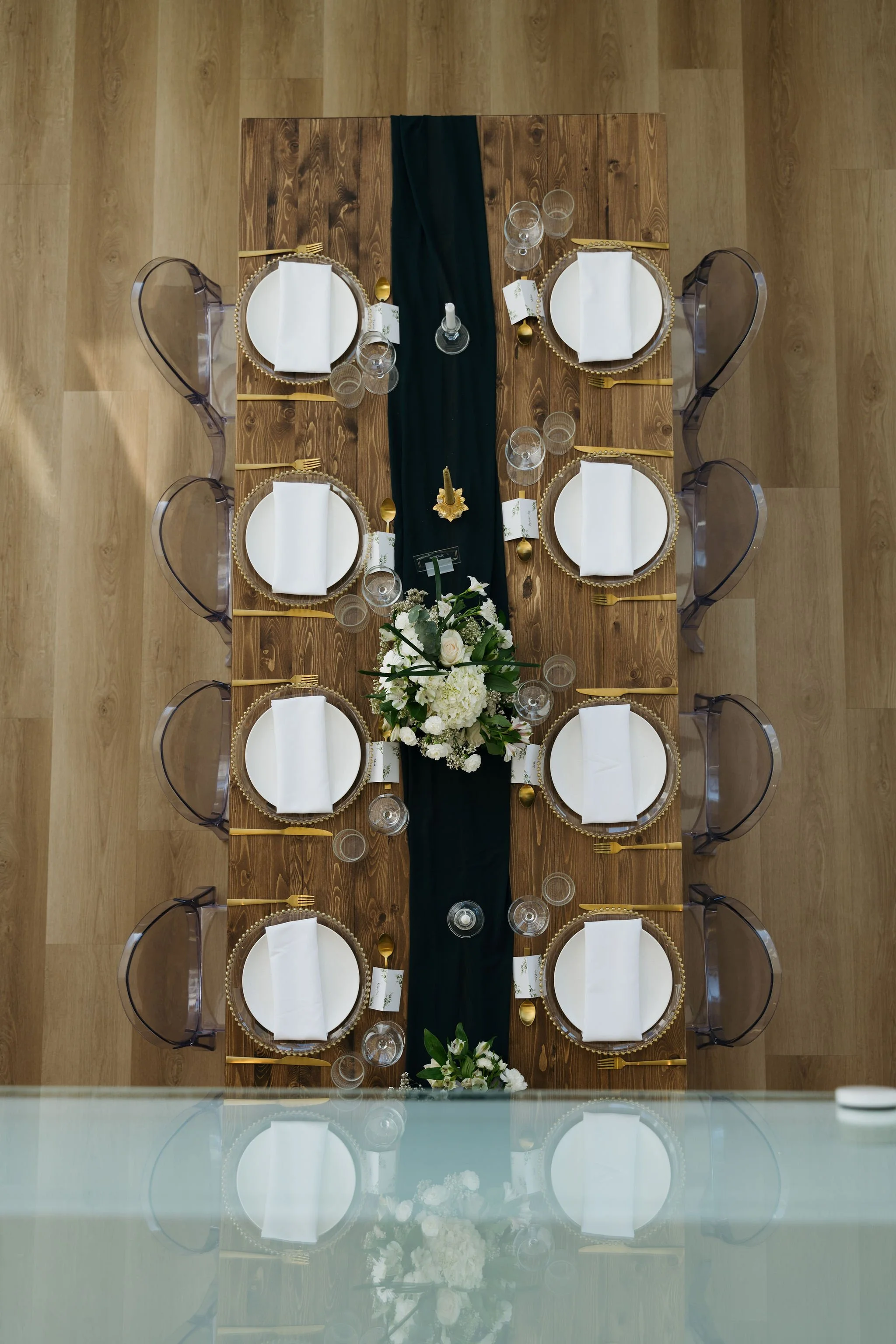Top-down view of elegant reception table styling at The Gem in Banff, with dark runner, gold-accent charger plates, crystal glassware, and a lush white floral centerpiece in a bright alpine-inspired space.