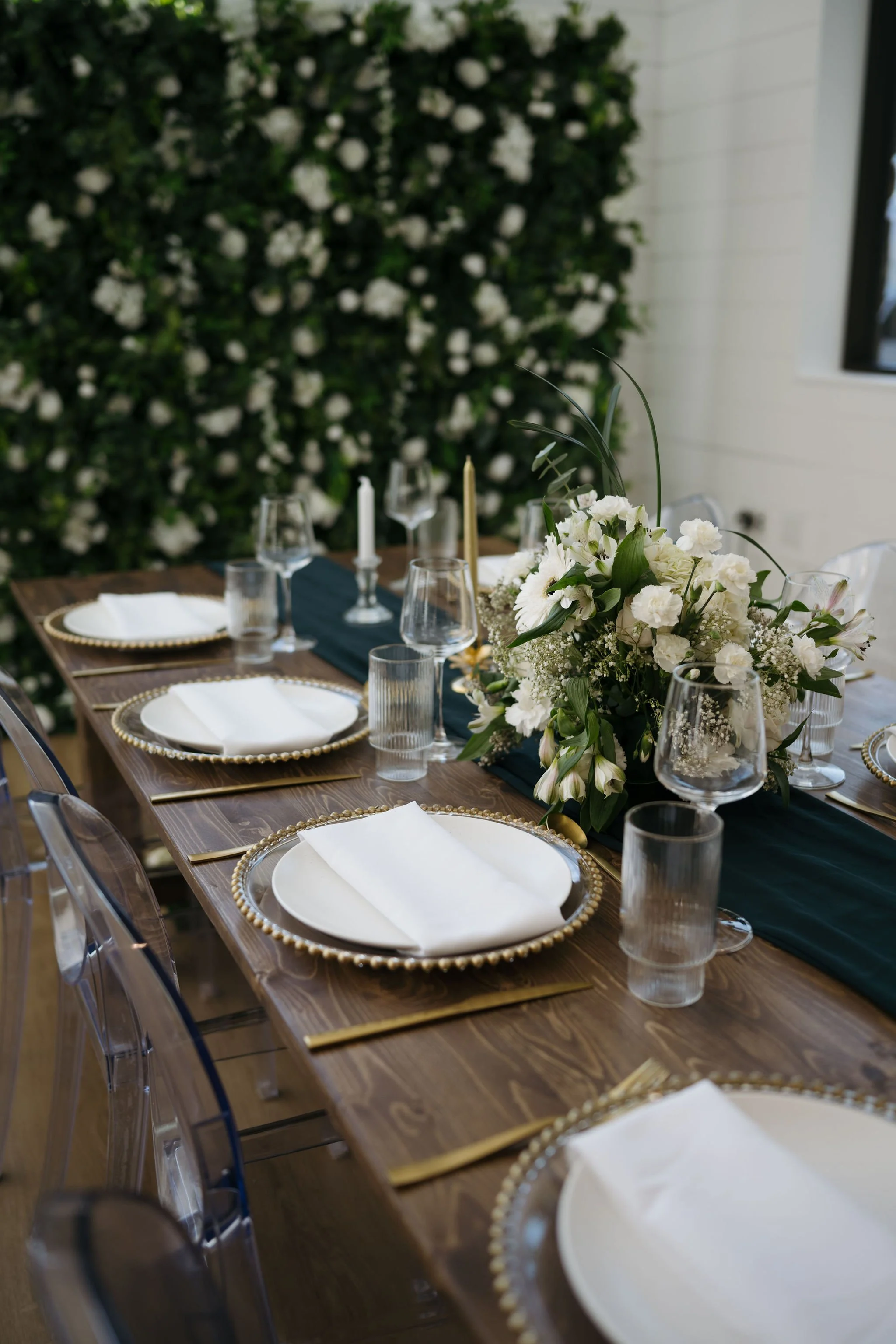 Focus on a floral centerpiece with a clear acrylic table number sign marked “Table 6” surrounded by white blooms, glassware, gold candleholders, and the edge of a dark runner.