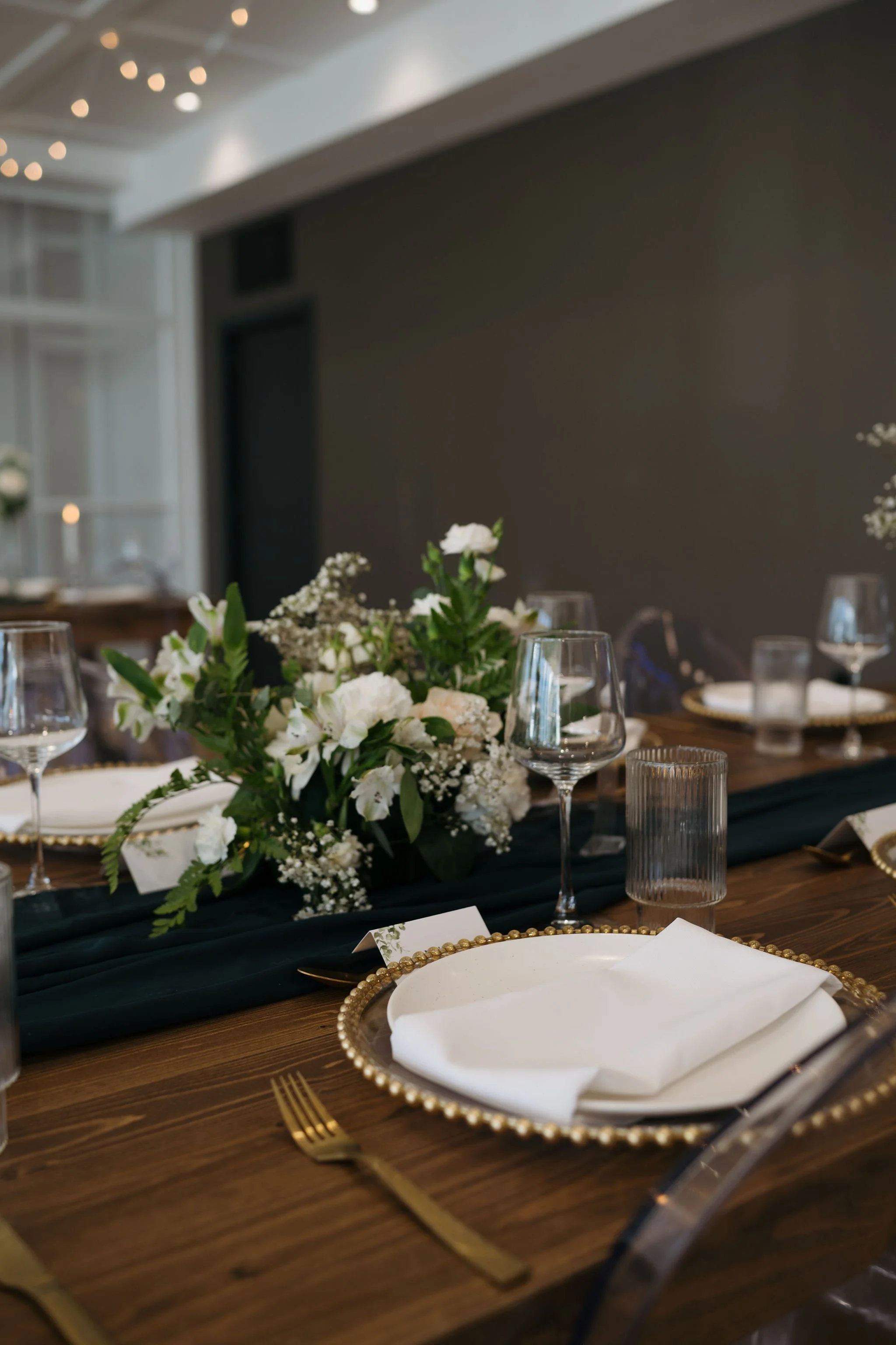 An elegant reception table is set with white plates framed by gold-beaded chargers, white cloth napkins, and modern gold cutlery. Glassware includes stemmed wine glasses and ribbed water glasses. A lush centerpiece of white flowers and greenery sits