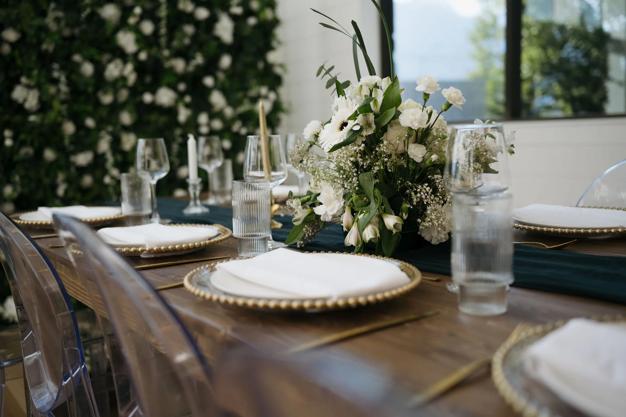 A deep wood table accented with a dark green runner, topped with lush white floral arrangements (carnations, gerbera daisies, baby's breath), candles and elegant glassware.