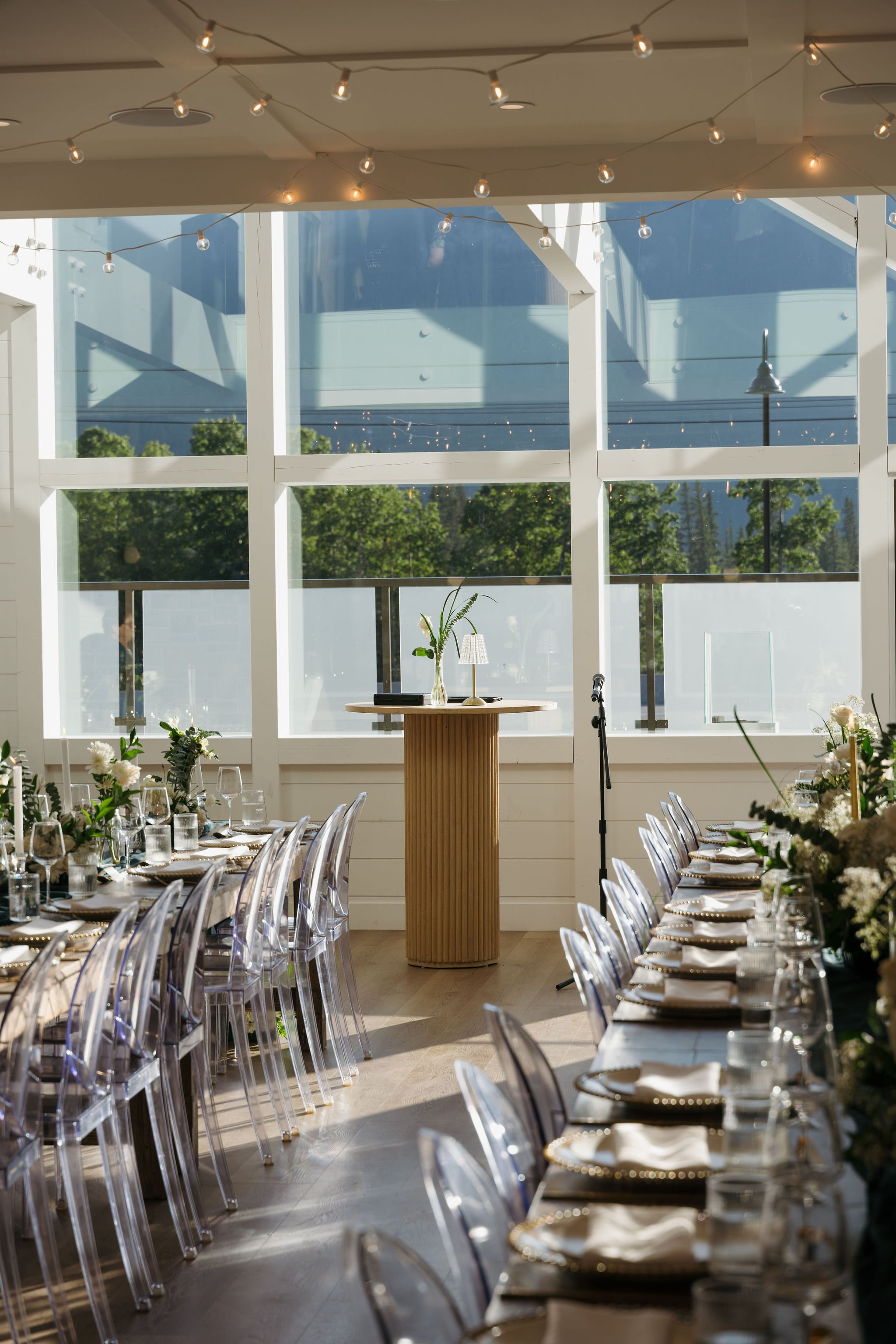 A bright indoor venue with floor-to-ceiling windows and hanging bistro string lights. Two long rows of tables with transparent ghost chairs lead to a tall round podium. The natural light and greenery visible outside enhance the fresh, open ambiance.