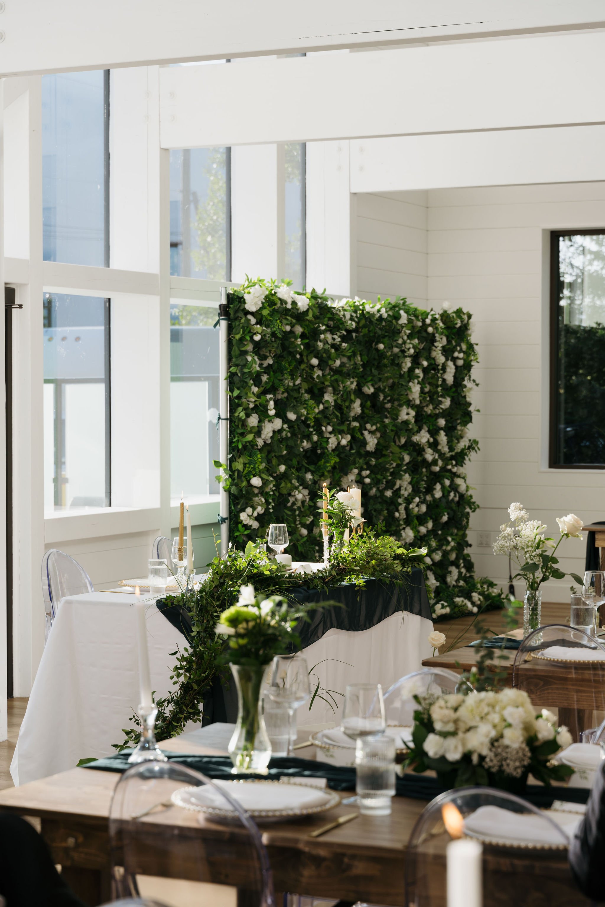 A sweetheart table draped in greenery and white fabric sits before a lush green floral backdrop. The table is styled with tall taper candles, delicate glassware, and minimalist white floral arrangements. Natural sunlight floods the space, emphasizing
