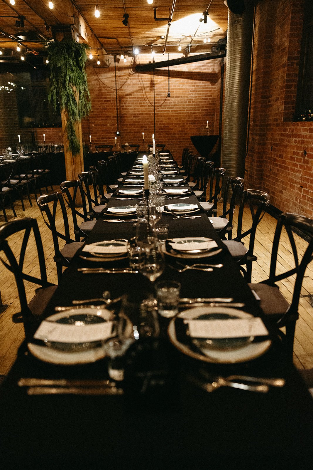 Moody reception interior with long banquet tables dressed in black linens, softly lit by candles and overhead string lights, featuring a central wood support column draped in greenery, linen seating, exposed brick walls and industrial ambiance.