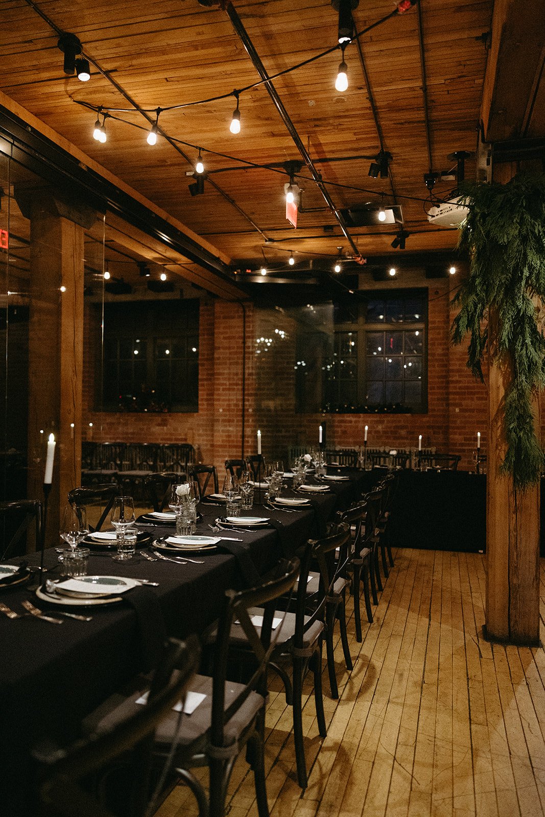 Moody reception interior with long banquet tables dressed in black linens, softly lit by candles and overhead string lights, featuring a central wood support column draped in greenery, linen seating, exposed brick walls and industrial ambiance.