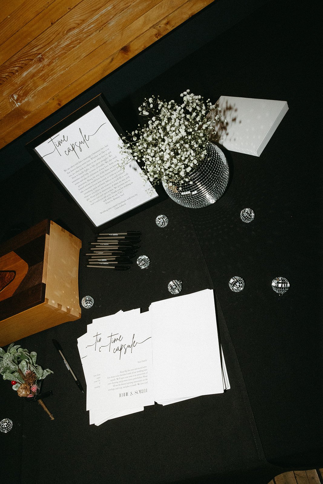 A guest table setup featuring a framed explanation titled Time Capsule, a mirrored vase of delicate white baby's breath, mini disco ball decor, pens, napkins, and printed cards for guests to write memories.