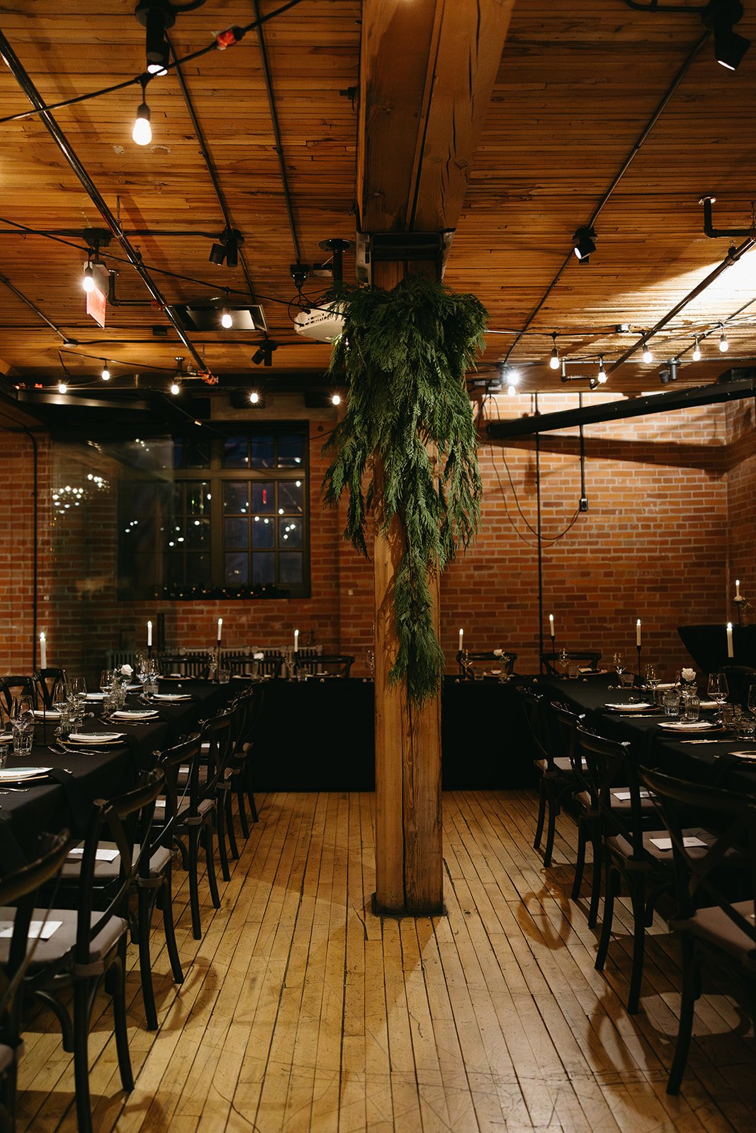 Moody reception interior with long banquet tables dressed in black linens, softly lit by candles and overhead string lights, featuring a central wood support column draped in greenery, linen seating, exposed brick walls and industrial ambiance.