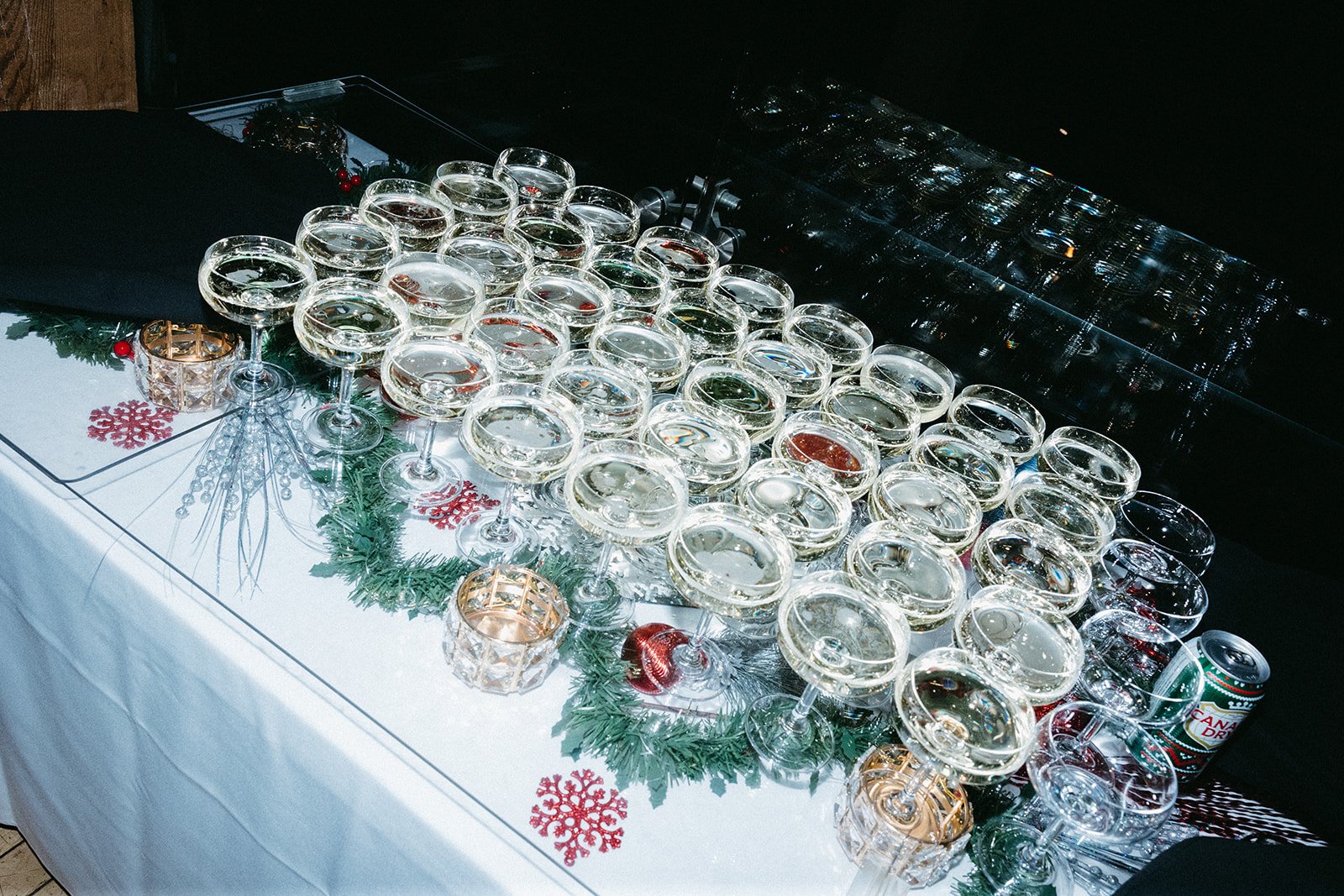 A shimmering display of coupe glasses filled with champagne, arranged in a tree-like shape on a table decorated with faux greenery, red ornaments, snowflake accents, and candle votives. A can of Canada Dry ginger ale sits nearby, adding to the celebr