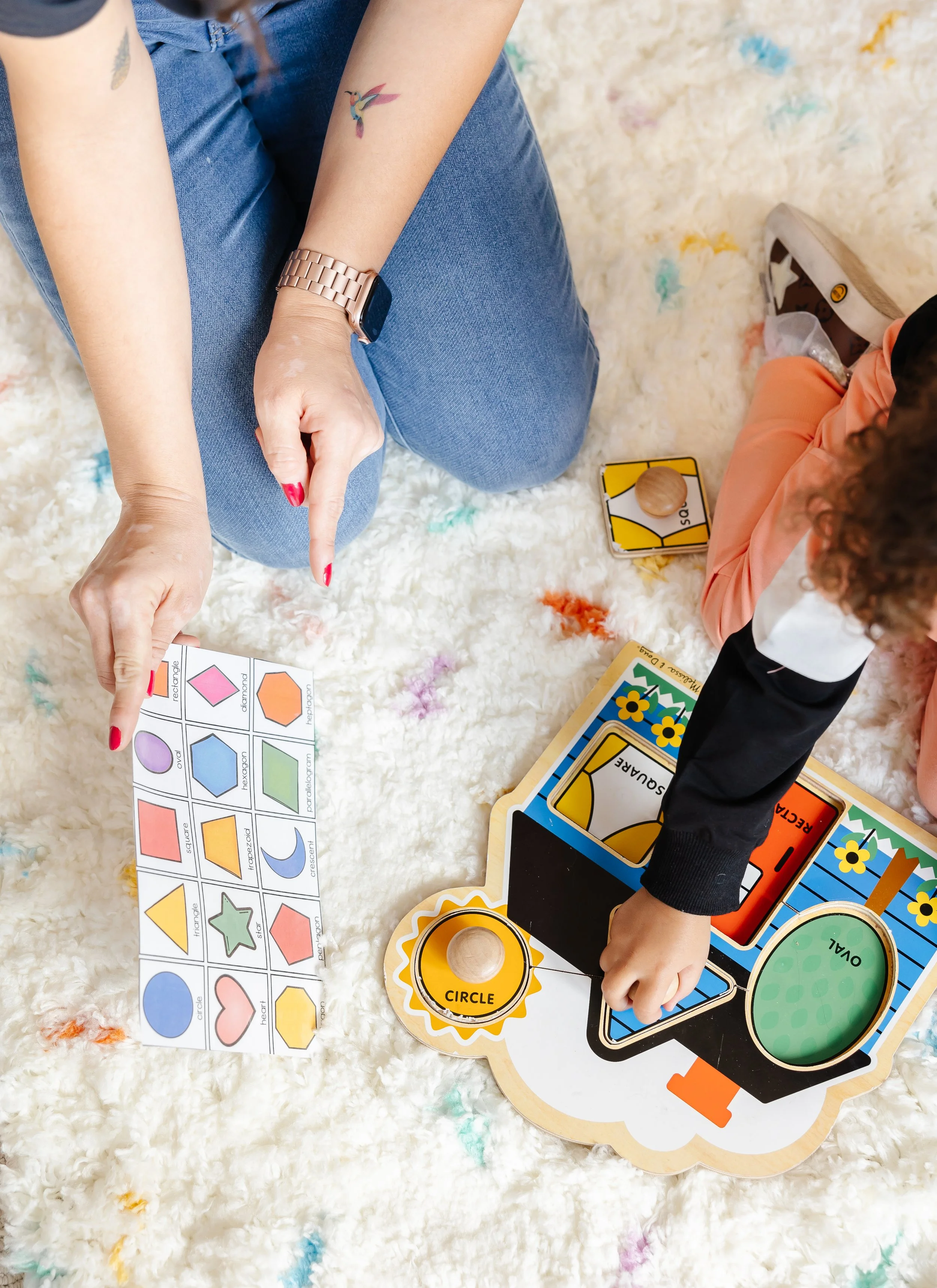 People playing an educational board game on a white, fluffy carpet with colorful confetti, involving shapes and colors.