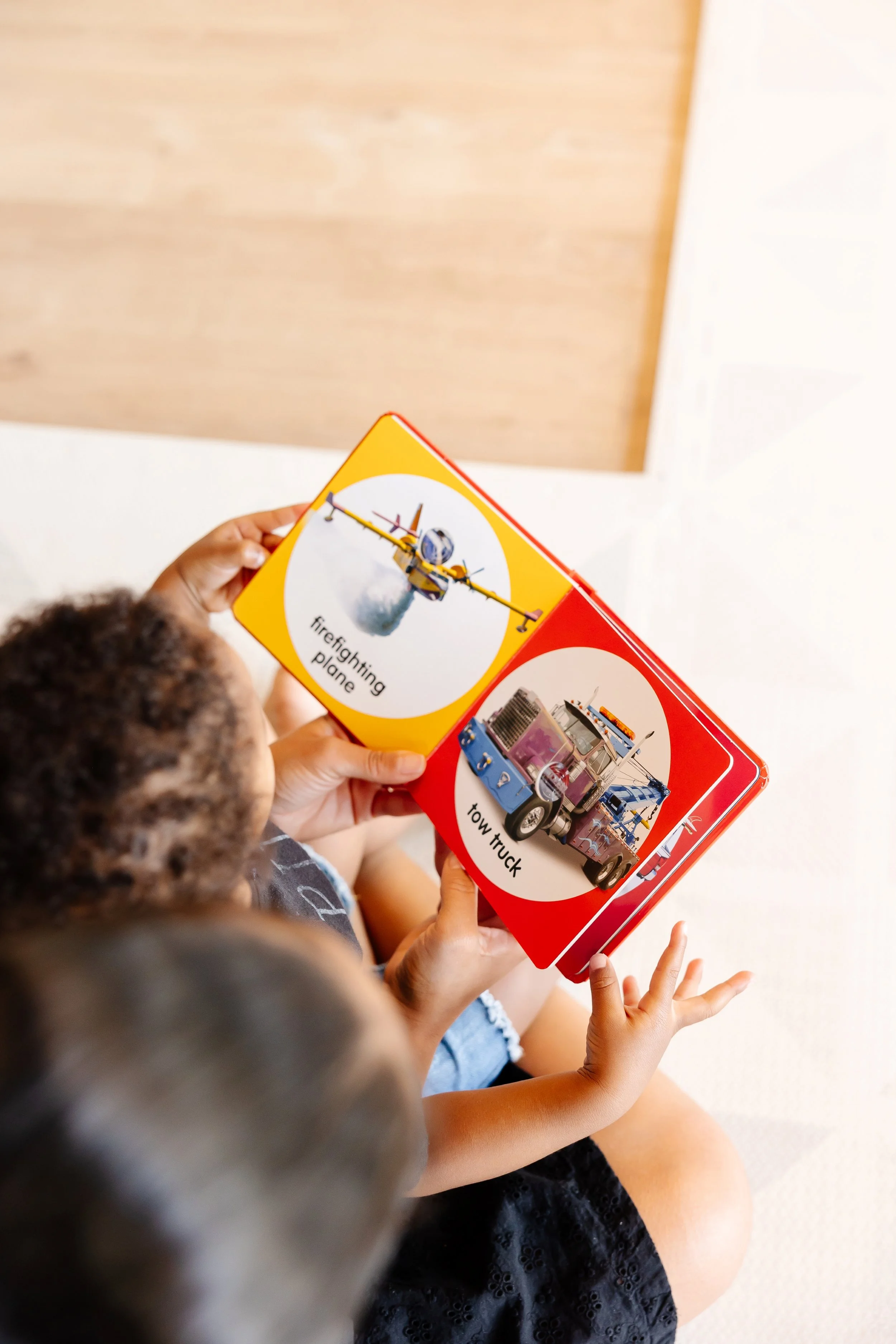 Two children reading a colorful picture book with pictures and labels of a firefighting airplane and a tow truck.