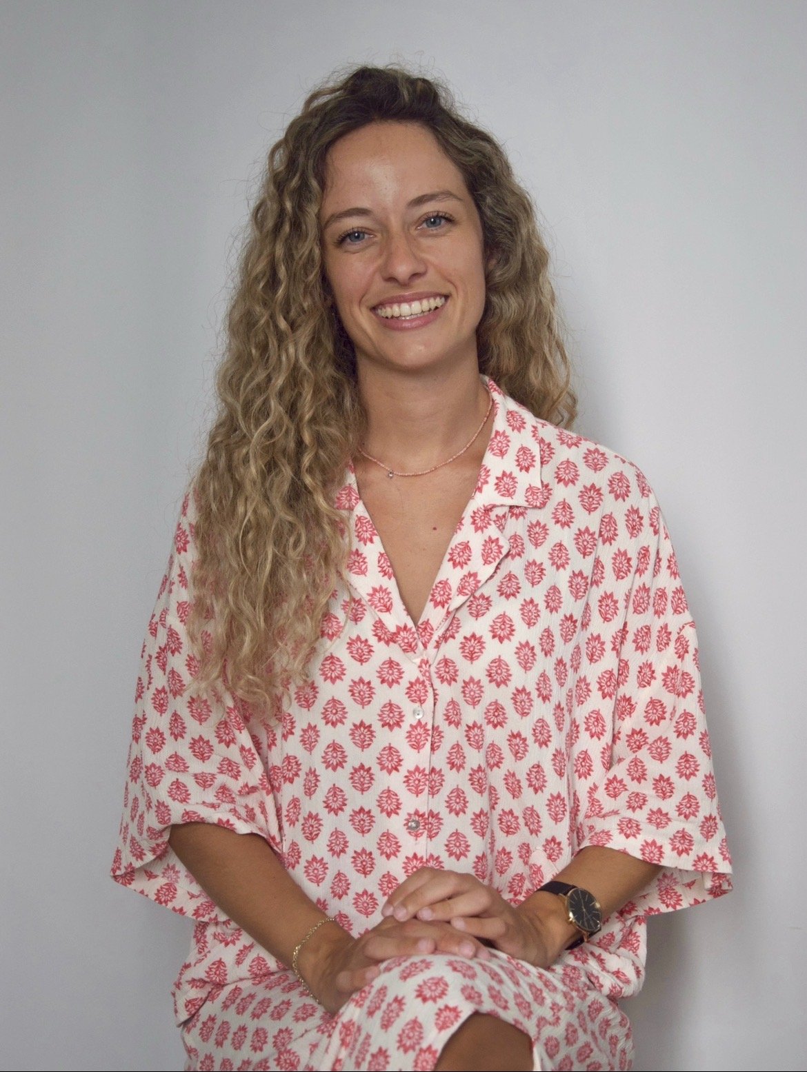 Mujer sonriendo con ropa rosa con patrón de flores, cabello rubio rizado, posando en pared blanca.