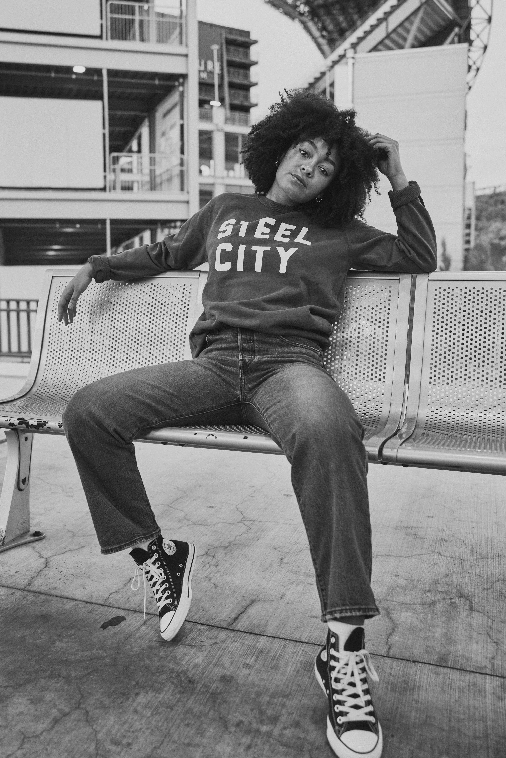 A woman with curly hair sitting on a metal bench in an urban setting, wearing a sweatshirt that says 'Steel City', jeans, and high-top sneakers, with modern buildings in the background.