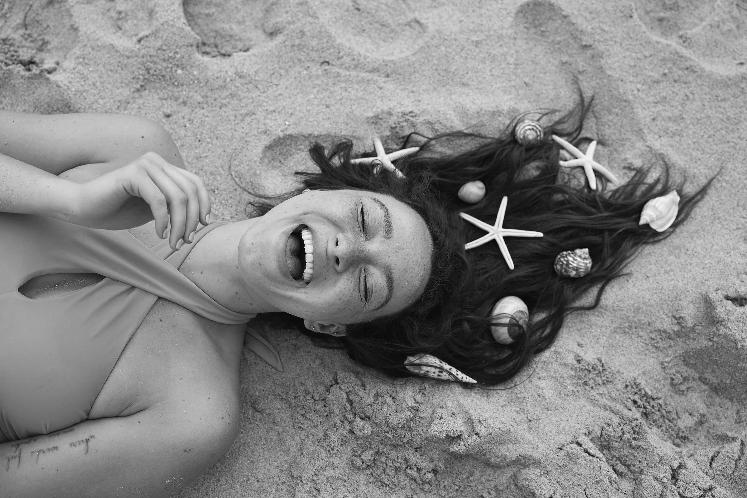 A young woman lying on the sand with her hair spread out, decorated with seashells and starfish, smiling and winking while holding a balloon.