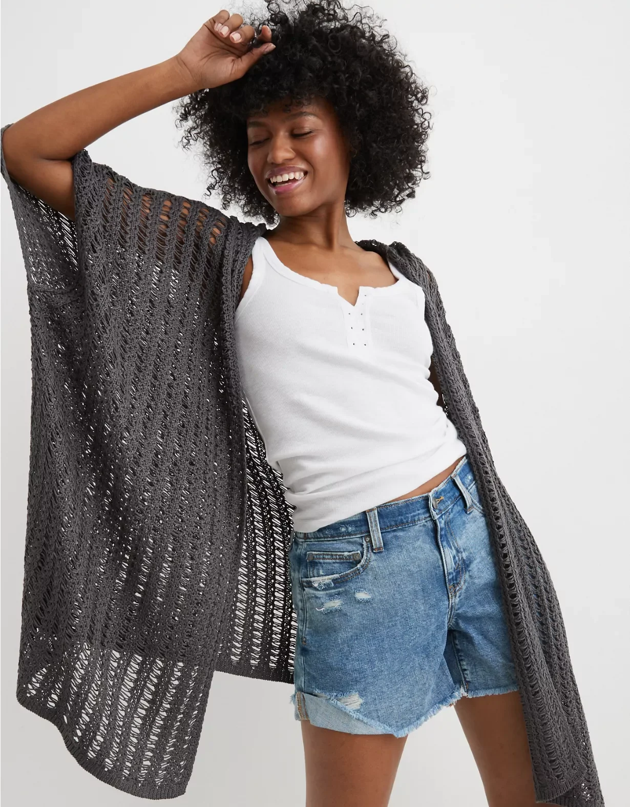 Woman in casual summer outfit posing with eyes closed and smiling, wearing a white tank top, distressed denim shorts, and a long gray open-knit cardigan.