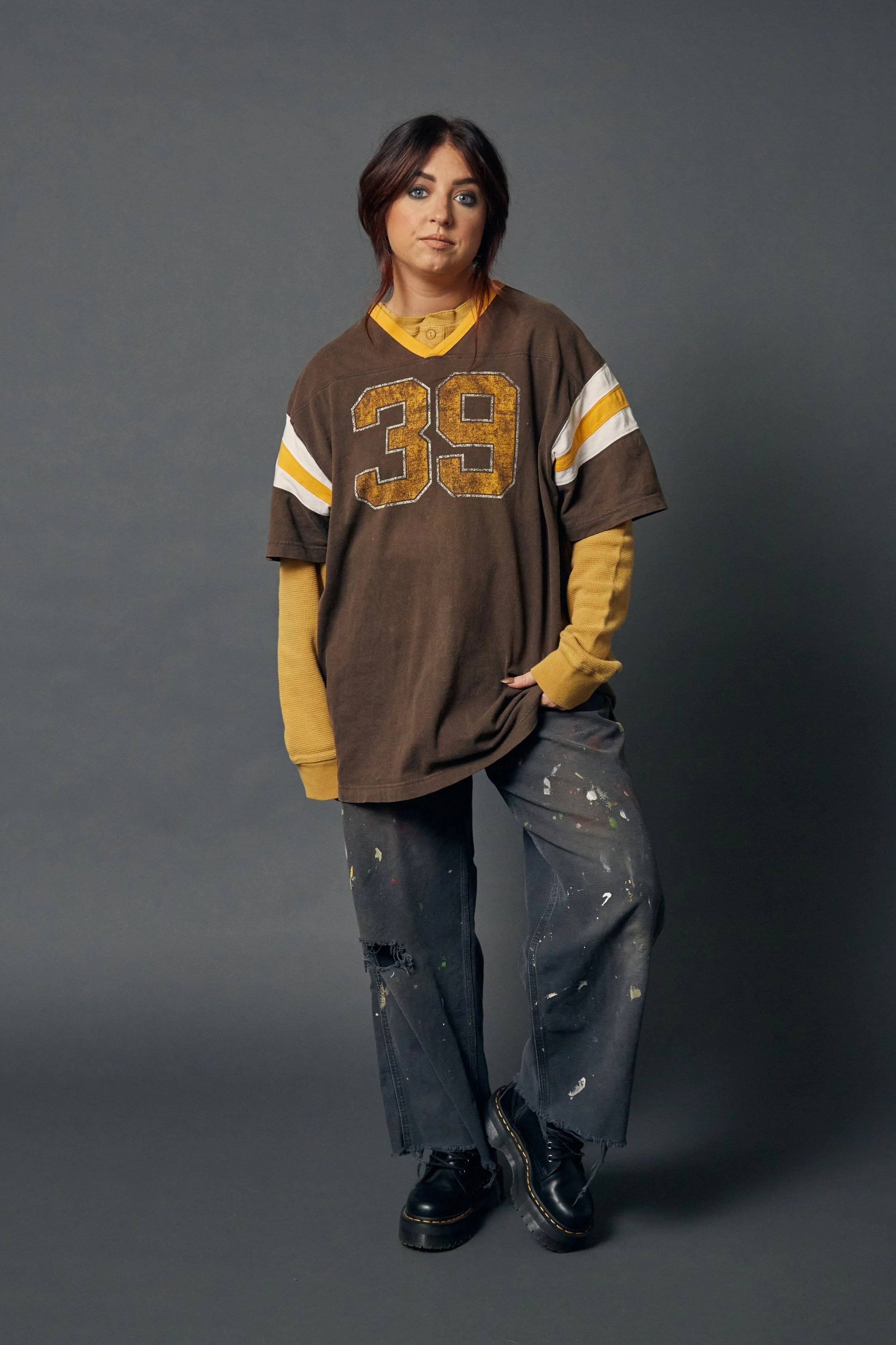 A woman with short brown hair and blue eyes standing in front of a gray background, wearing a oversized brown sports jersey with the number 39, yellow long-sleeve shirt underneath, distressed black pants with paint splatters, and black combat boots.