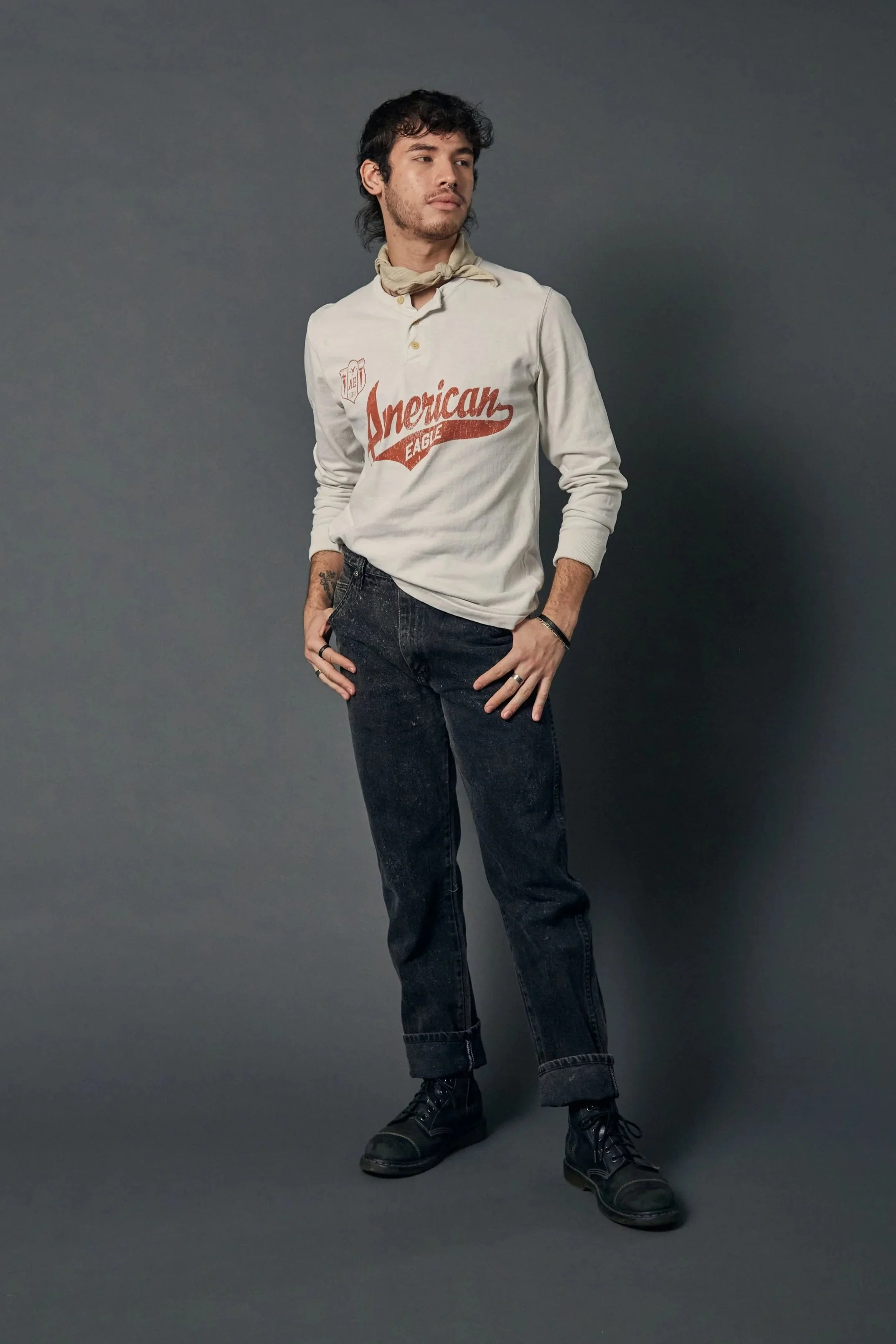 A young man with dark, curly hair stands against a gray background, wearing a beige long-sleeve shirt with red text that reads 'American Eagle,' black jeans, black boots, and has a beige bandana tied around his neck.