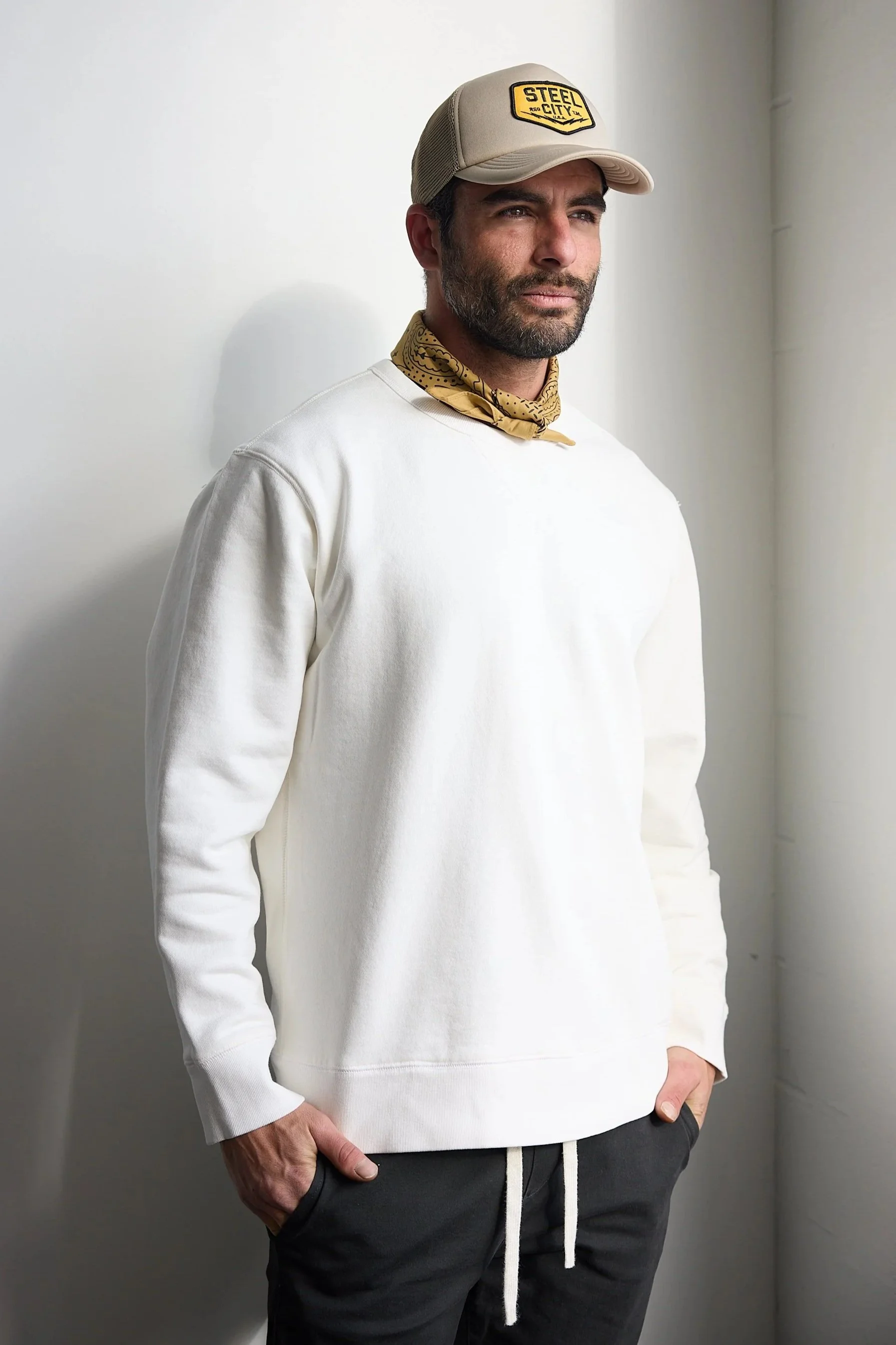 A man wearing a beige cap, a yellow bandana, a white sweatshirt, and black pants standing against a white wall.