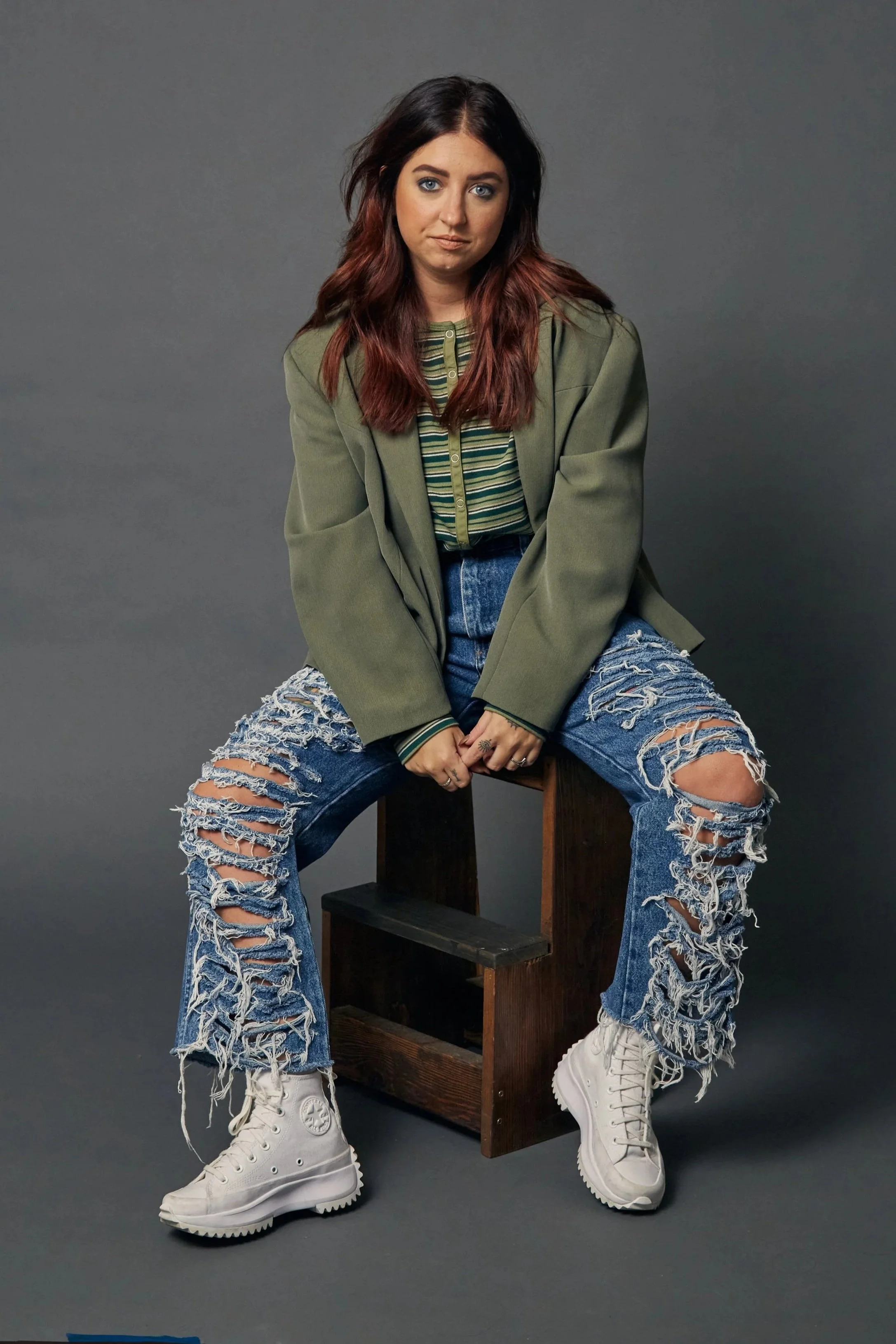 A young woman with long wavy brown and red hair, wearing an oversized green jacket, ripped blue jeans, a striped shirt, and white high-top sneakers, sitting on a wooden stool against a gray background.