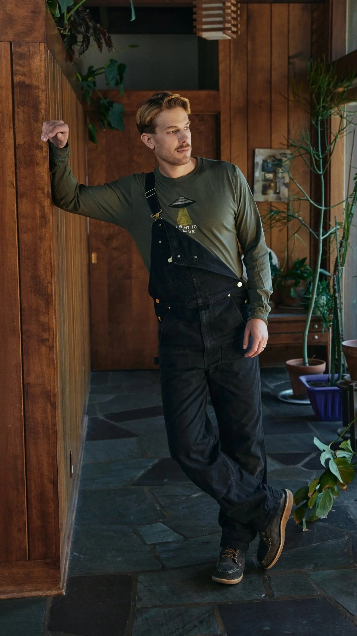A young man with blond hair and a beard standing inside a room with wood-paneled walls, leaning against a wall with his right arm, wearing a green long-sleeve shirt, black overalls, and black boots, looking out of a window with indoor plants around.
