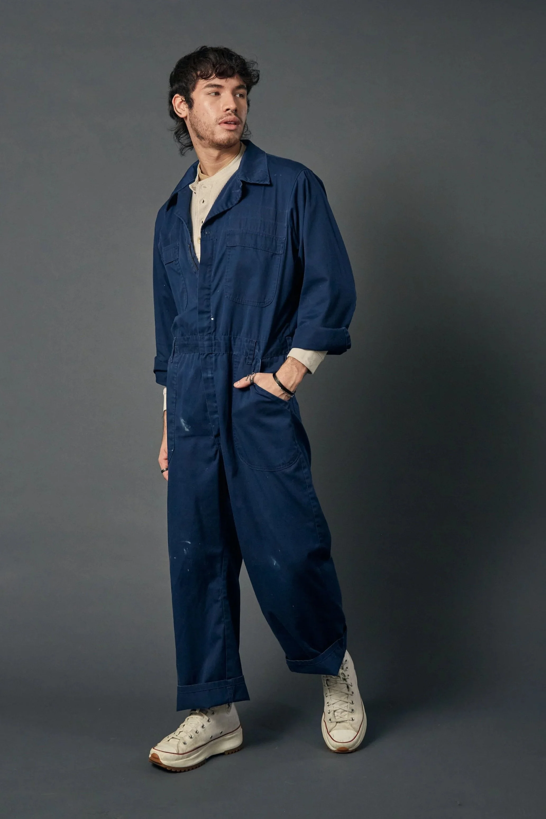 A young man with curly dark hair is standing against a gray background, wearing a navy blue jumpsuit and off-white high-top sneakers.