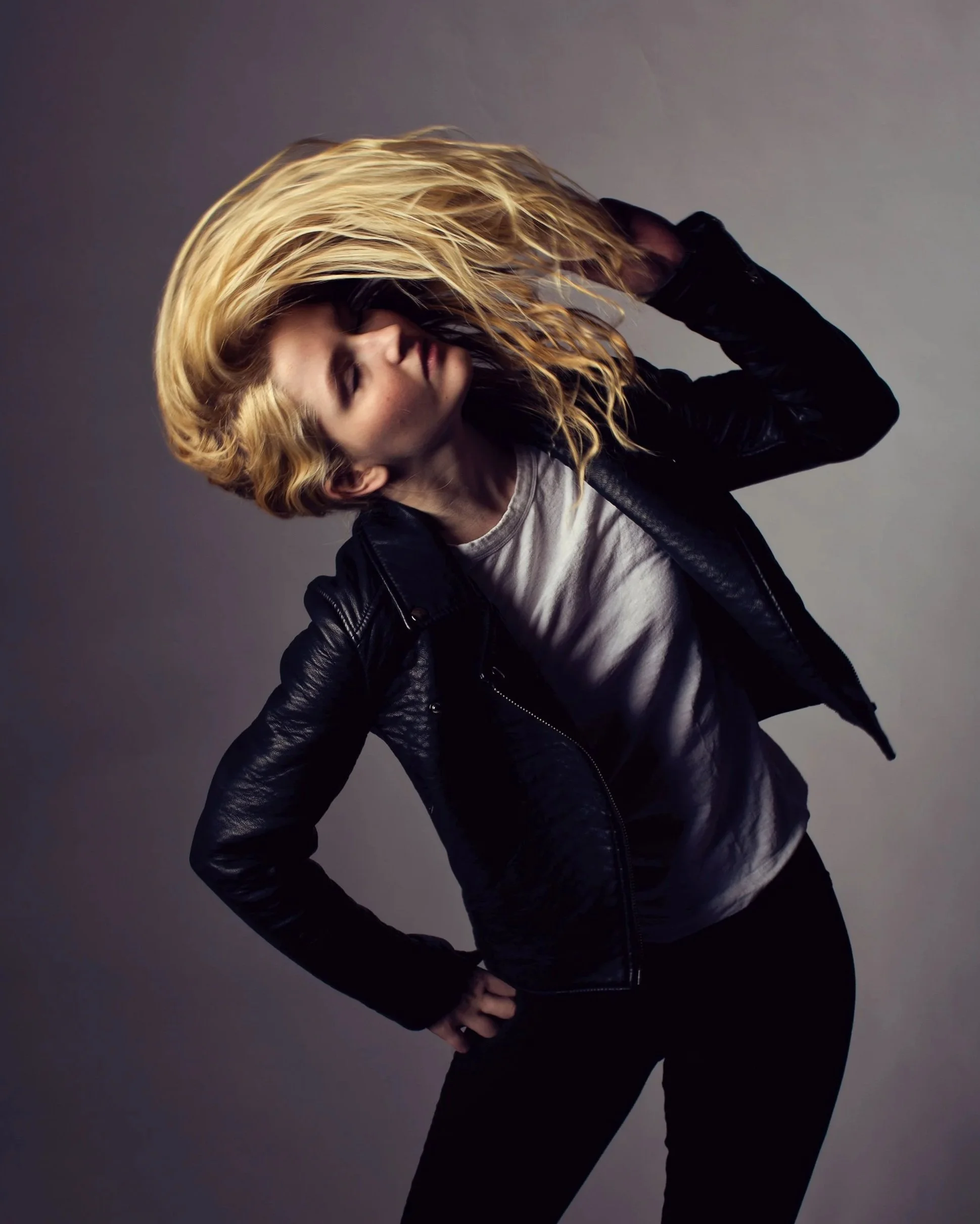 Person with long blonde hair, wearing a black leather jacket and white shirt, flipping hair against a gray background.