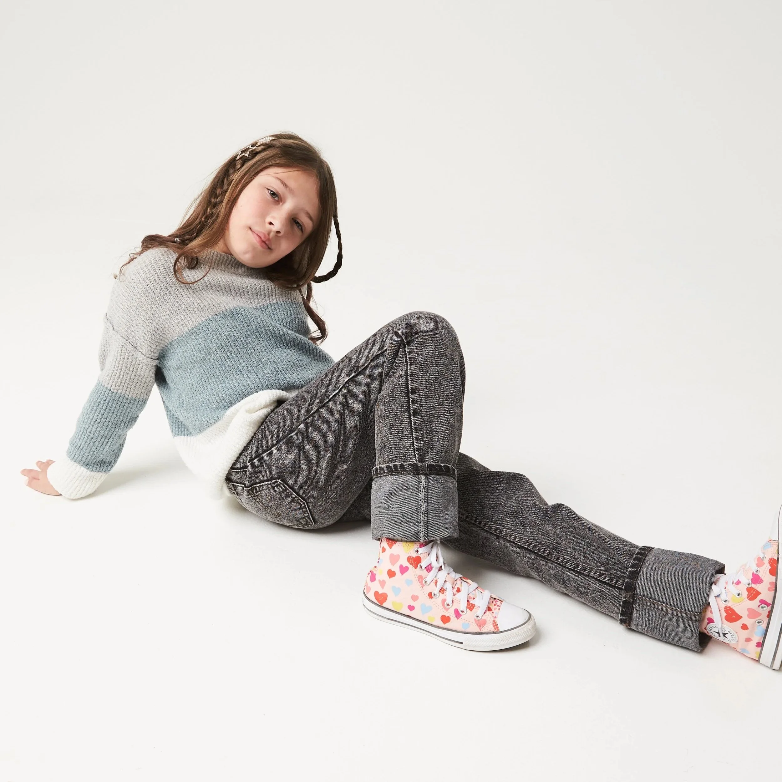 A young girl with brown hair wearing a multicolored knit sweater, dark gray jeans, and colorful high-top sneakers, sitting on a white background.