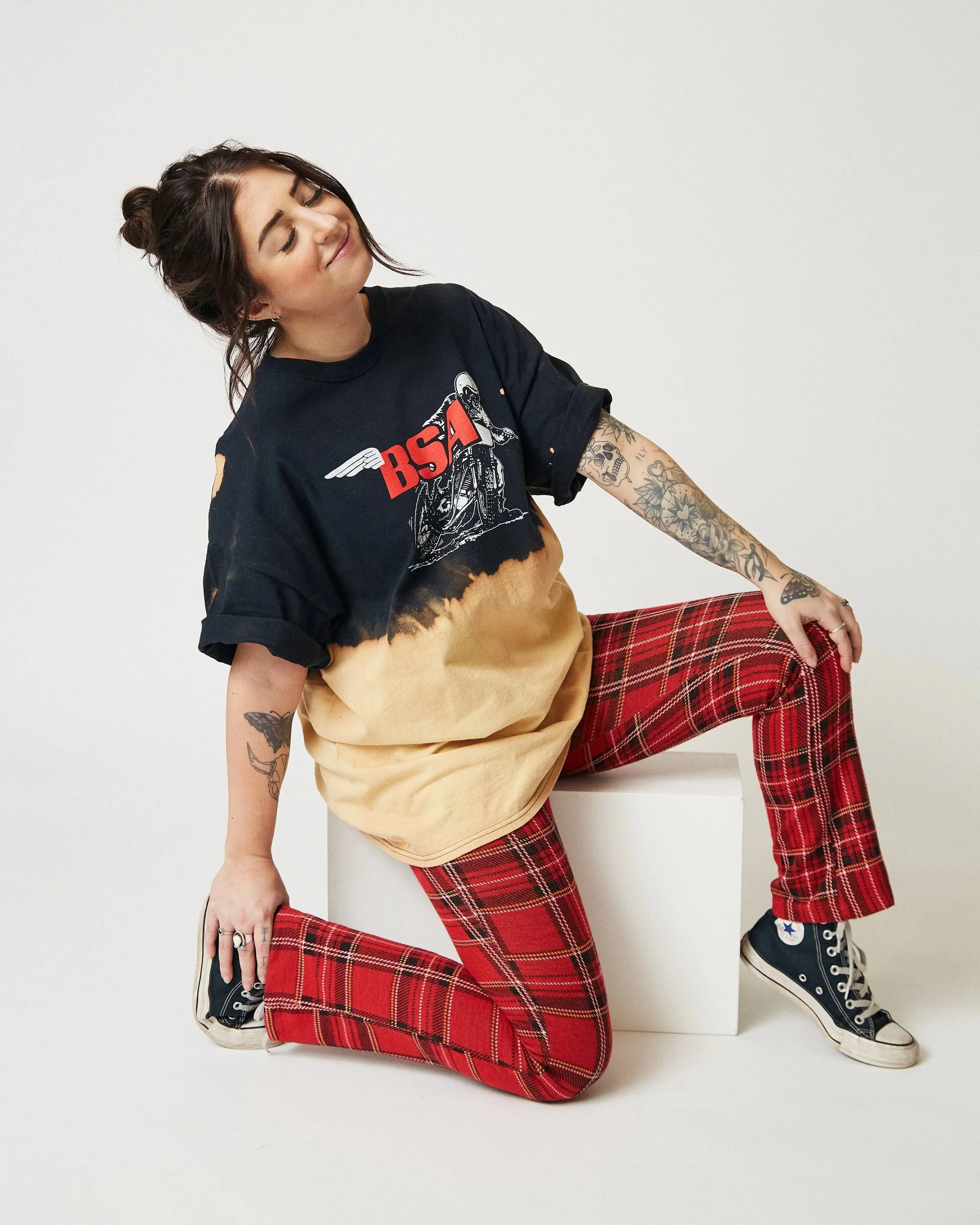 A woman with dark hair in a bun, tattoos on her arms, wearing a vintage black and tie-dye t-shirt, red plaid pants, and black high-top sneakers, posing in a kneeling position on a white surface against a plain white background.