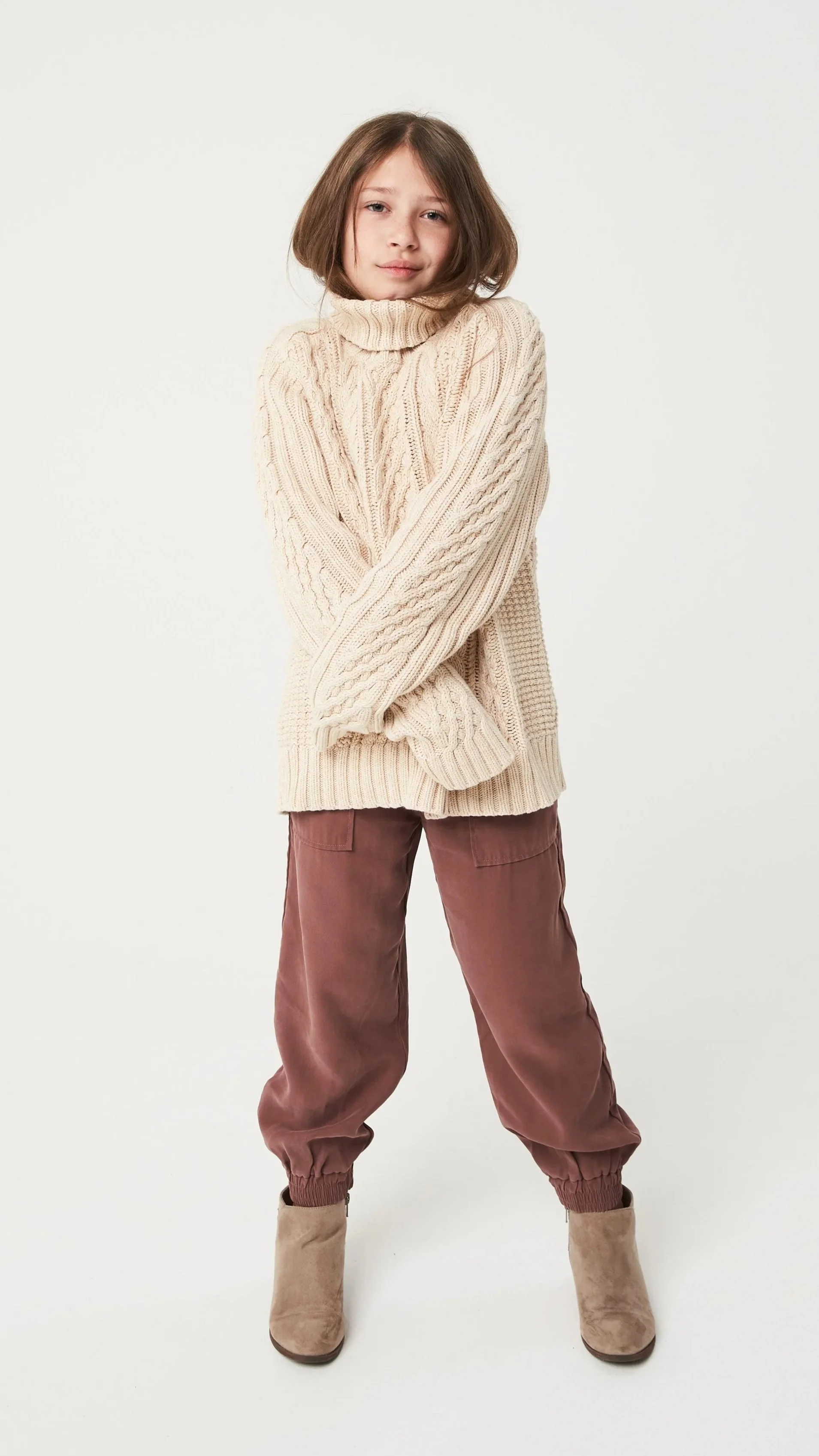 Young girl with shoulder-length brown hair wearing a cream cable-knit sweater, brown pants, and beige ankle boots standing against a plain white background