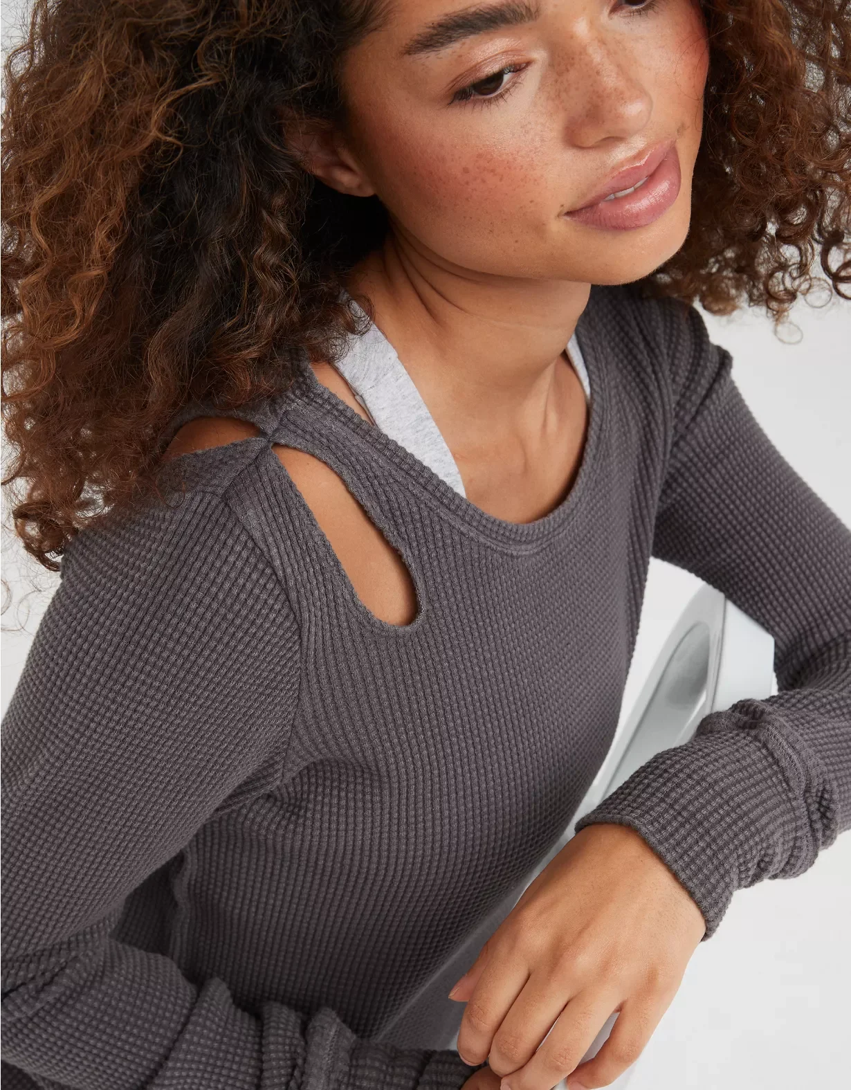 Close-up of a woman with curly hair and freckles wearing a gray sweater with shoulder cutouts and a gray sports bra underneath.