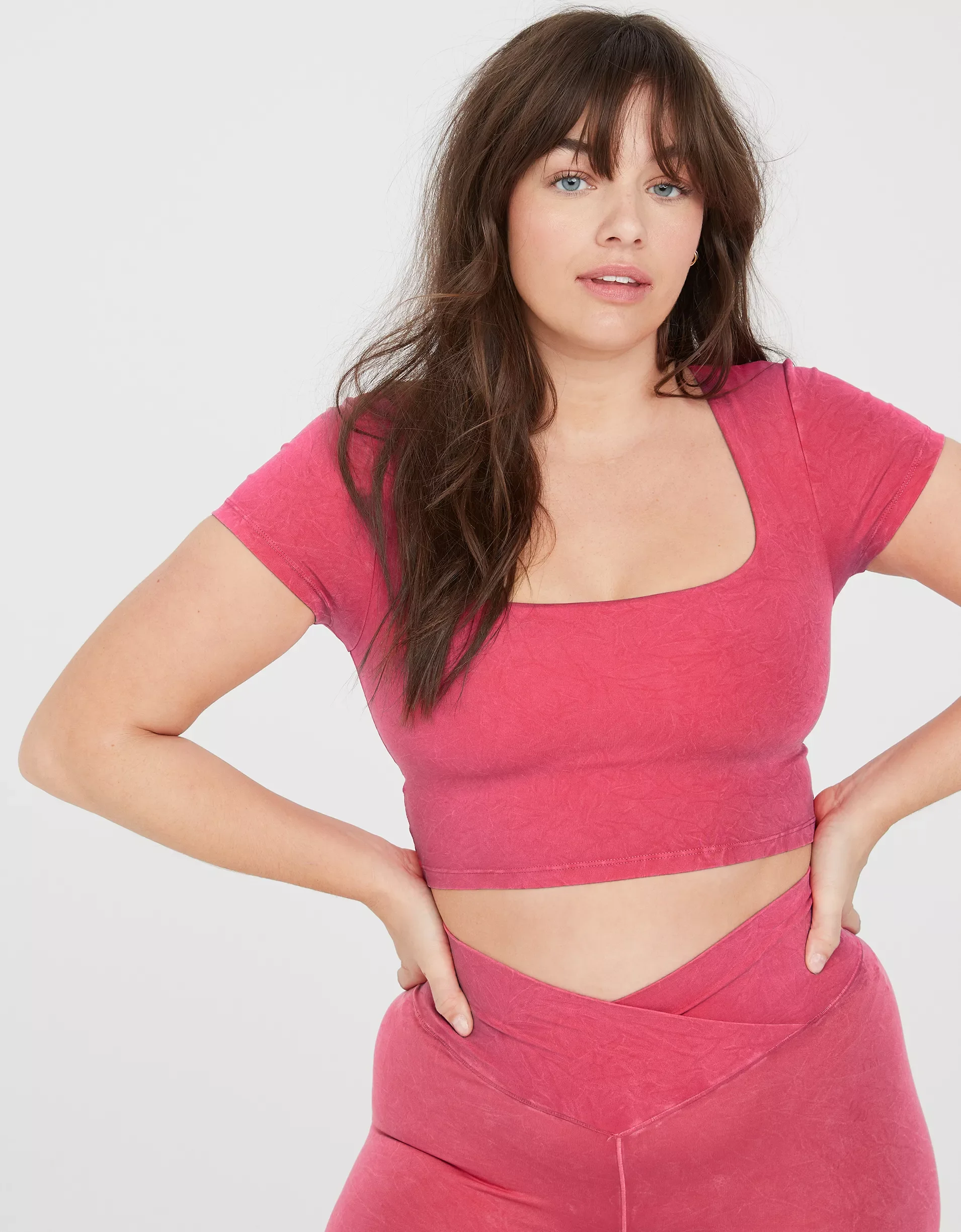 Woman with long brown hair, blue eyes, wearing a pink crop top and matching workout leggings, standing with hands on hips against white background.
