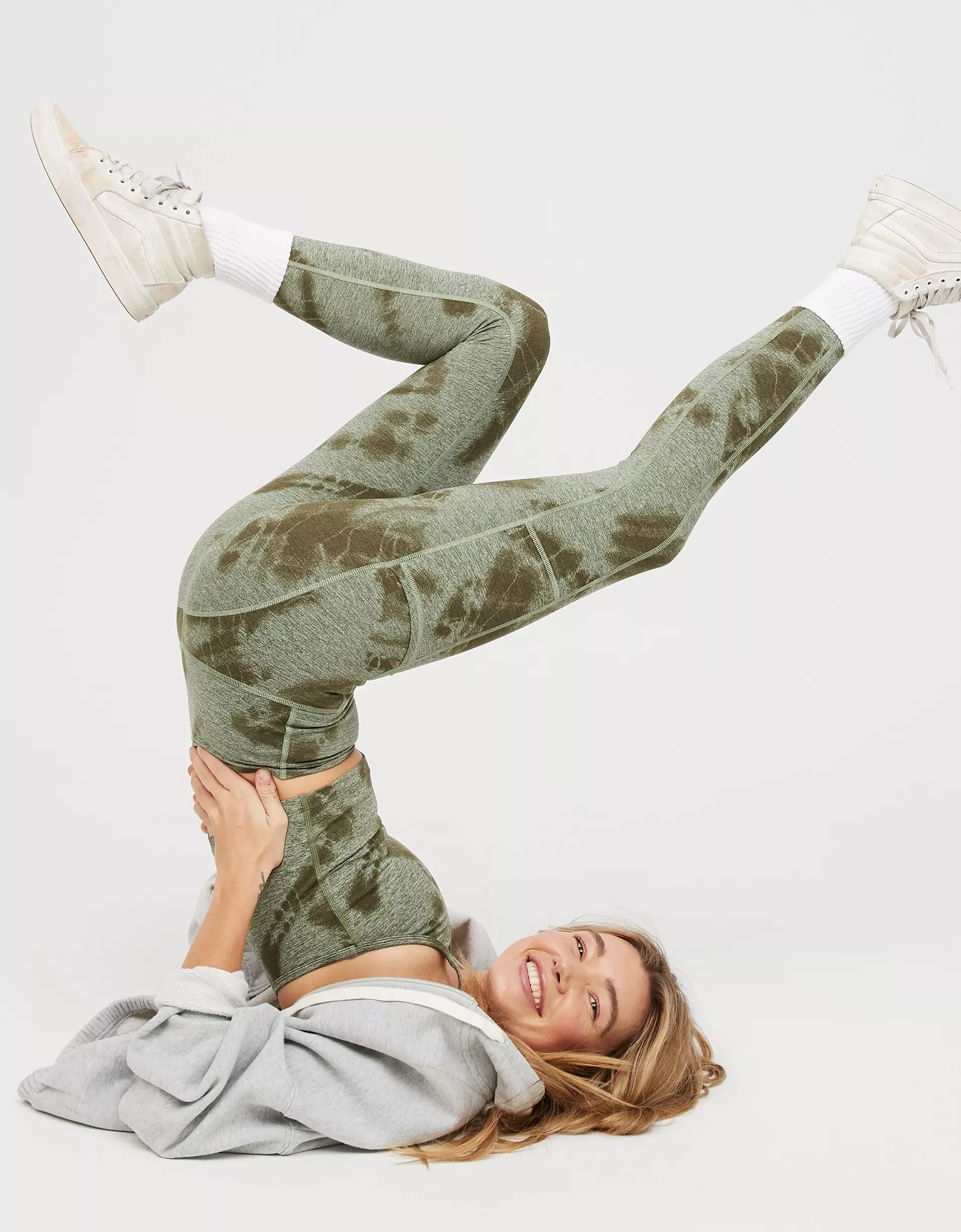 A woman with long hair is lying on her back on a plain white background, smiling while holding her legs in the air.