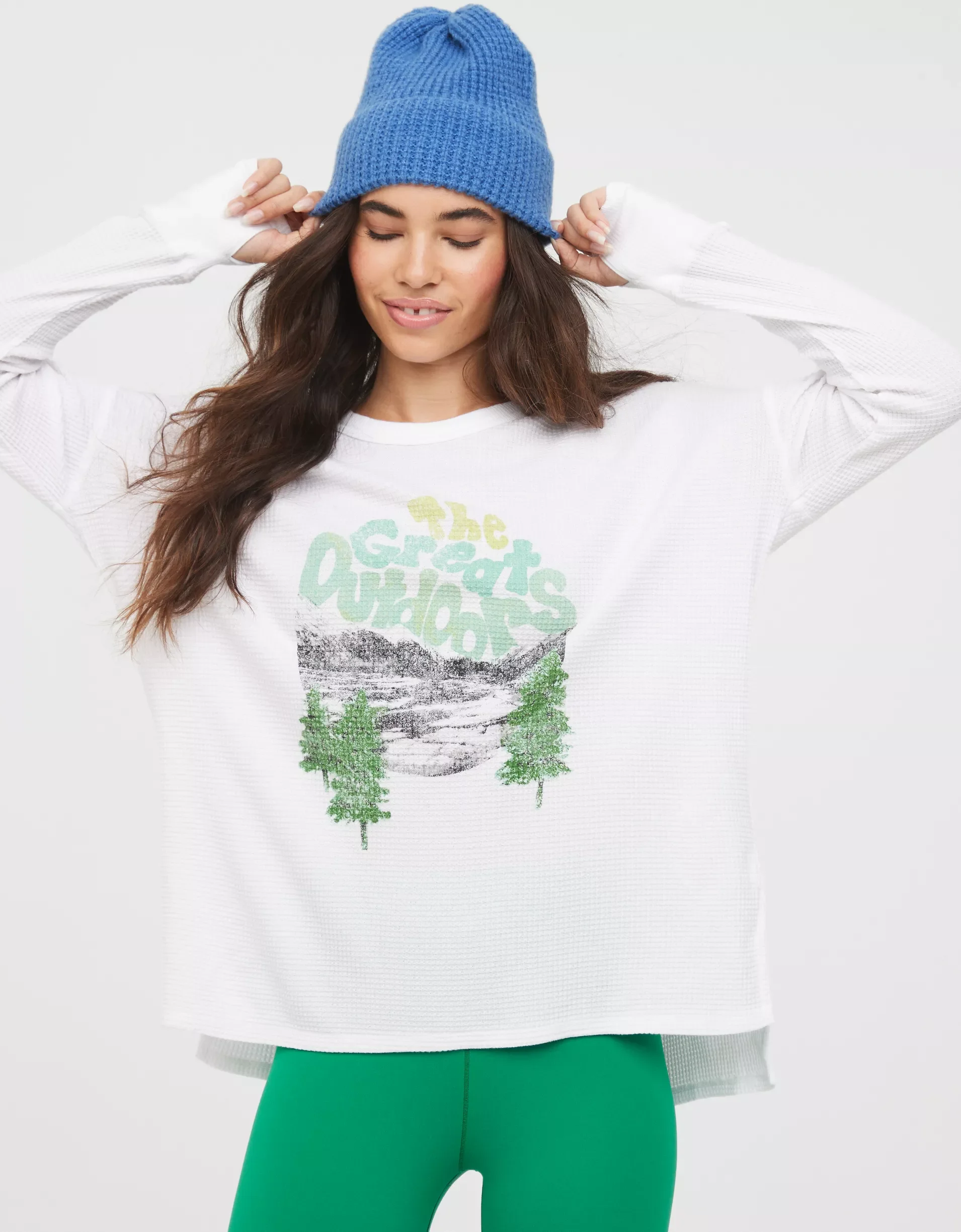 Woman wearing a blue knit beanie, white long-sleeve shirt with graphic of mountains, trees and text that reads 'The Great Outdoors', and green leggings.